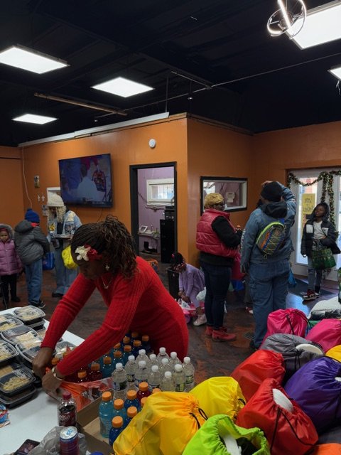People gathering in a community center with tables of bottled drinks and bags, some people waiting in line, some looking at food, in an indoor space with orange walls and ceiling lights.