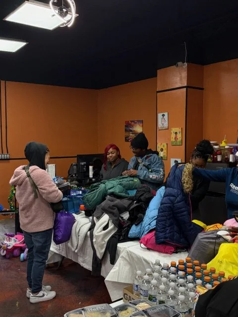 People at a table in a room, possibly in a community center, with various items and snacks on the table and in the background.