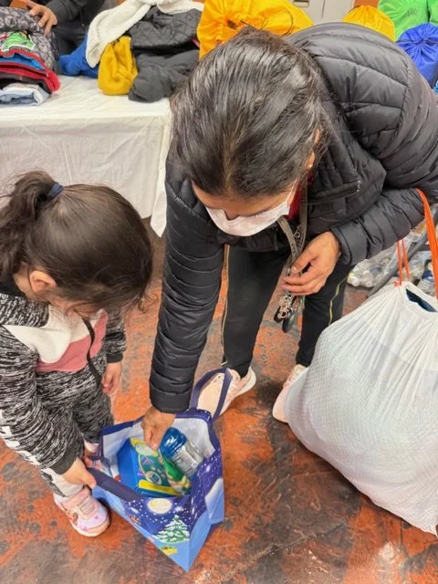 Woman and young girl packing clothing and essentials into a holiday-themed gift bag surrounded by more clothing and bags.