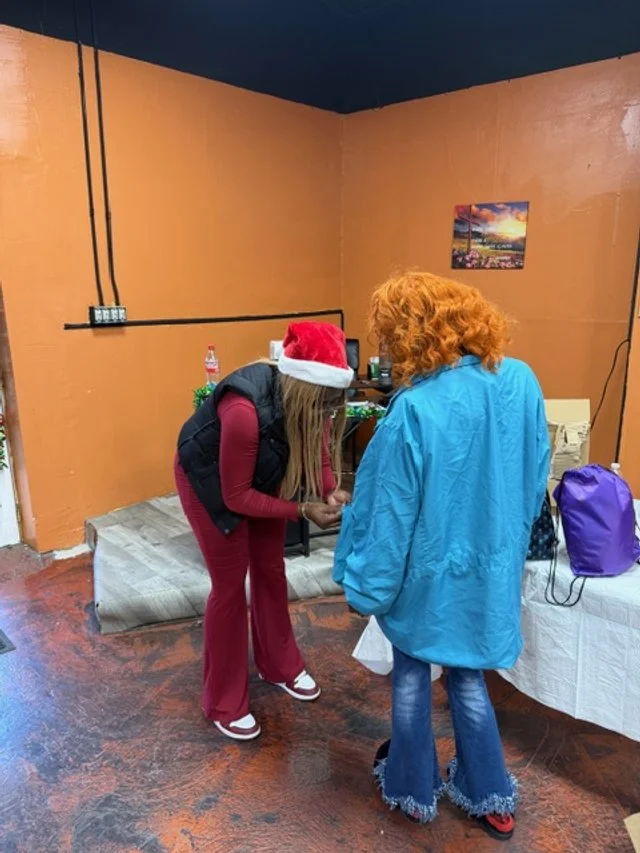 Two women, one wearing a Santa hat and the other with curly red hair, are standing and talking to each other in a room with orange walls. The room has a table with a purple bag and some electronics on it.