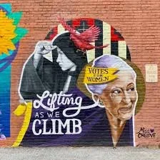 A mural featuring women climbers with the text 'Lifting As We Climb' and a woman holding a sign that says 'Votes for Women,' advocating for women's independence and suffrage.