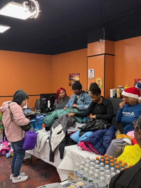 Group of people sorting through clothes on a table, with shopping bags, water bottles, and a child in a Santa hat nearby, in a room with orange walls.