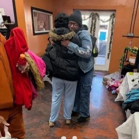 Two people hugging inside a room, with one wearing a black beanie and jacket, and the other in a gray hoodie.