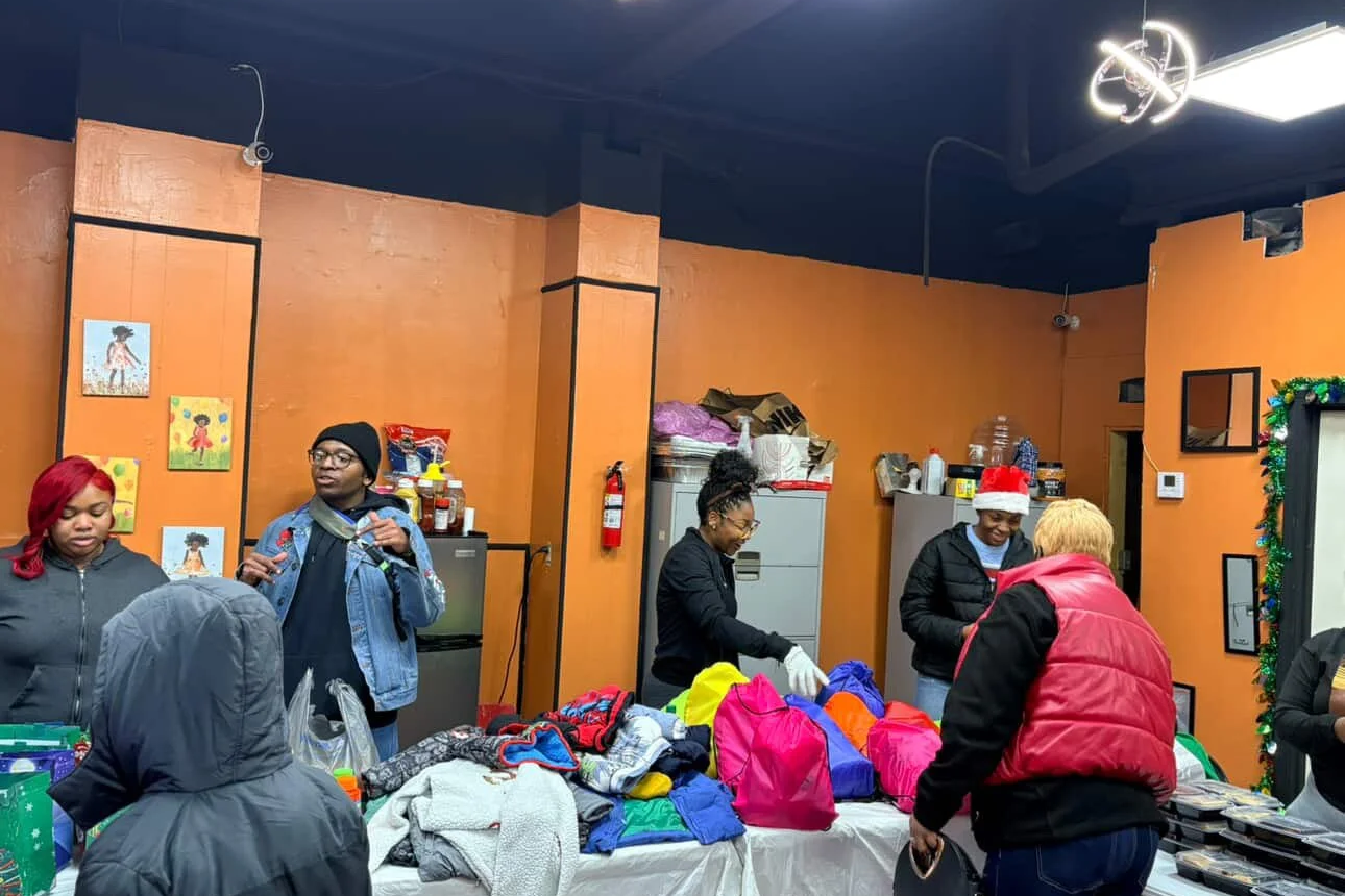 People gathered around a table with colorful bags and clothing, some wearing holiday hats, indoors with orange walls decorated with children's artwork.