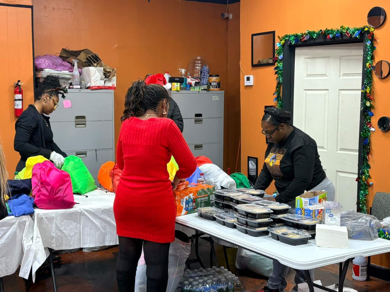 People preparing or distributing food in a room decorated with Christmas garlands. Several containers of food are on a table, and other supplies are being organized.