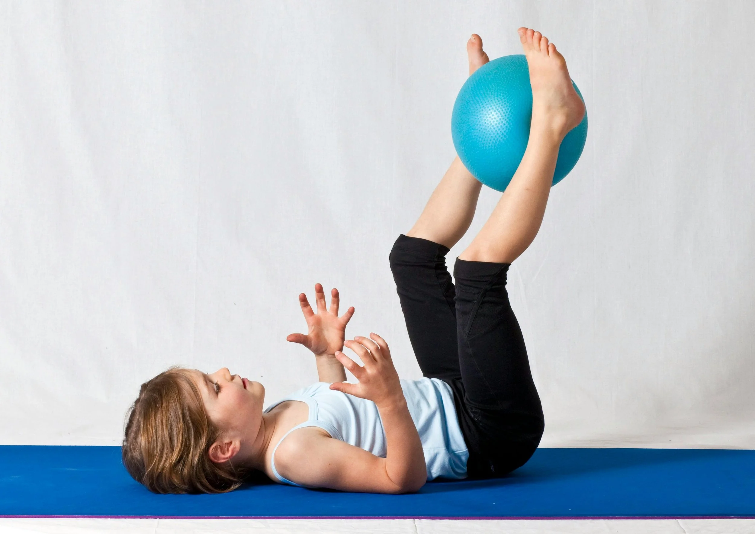 A young girl lying on her back on a blue exercise mat with her legs raised in the air, holding a blue exercise ball between her knees, with her arms bent and hands open.