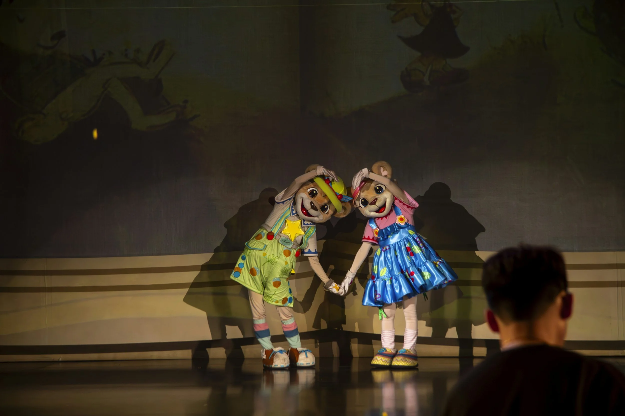 Two performers in colorful Disney character costumes holding hands on stage with a dark background and a person watching in the foreground.