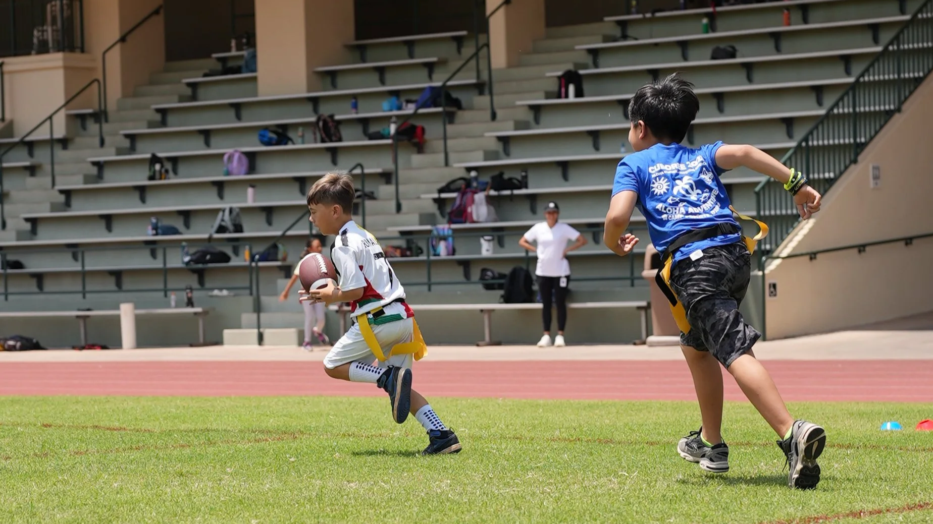 Two boys playing football on a field with bleachers in the background. One boy is running with the ball, and another is chasing him.