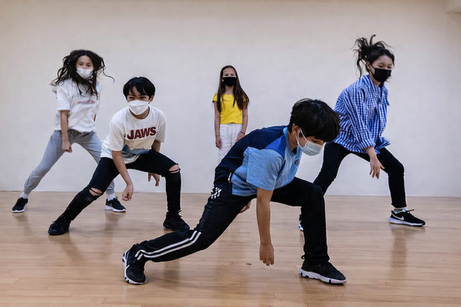 Five children practicing dance moves in a studio, all wearing face masks and casual clothing.