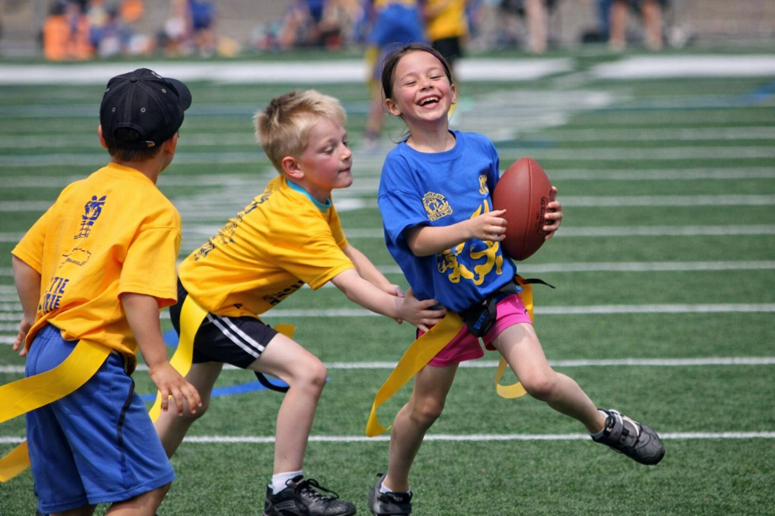 Children playing flag football on a sports field, one girl in a blue shirt holding a football and laughing, two boys in yellow shirts trying to pull her flag.
