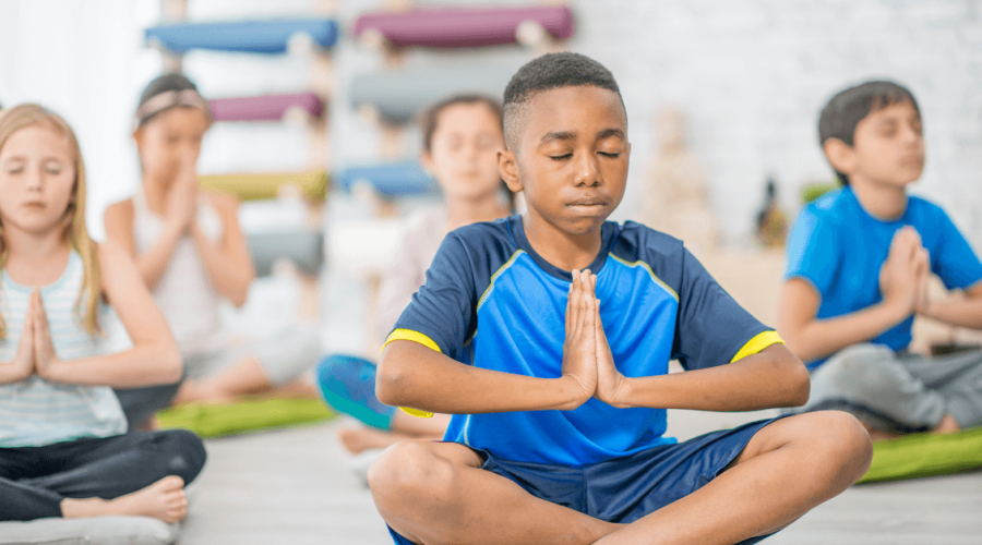 Children meditating in a yoga class, sitting cross-legged with hands in prayer position.