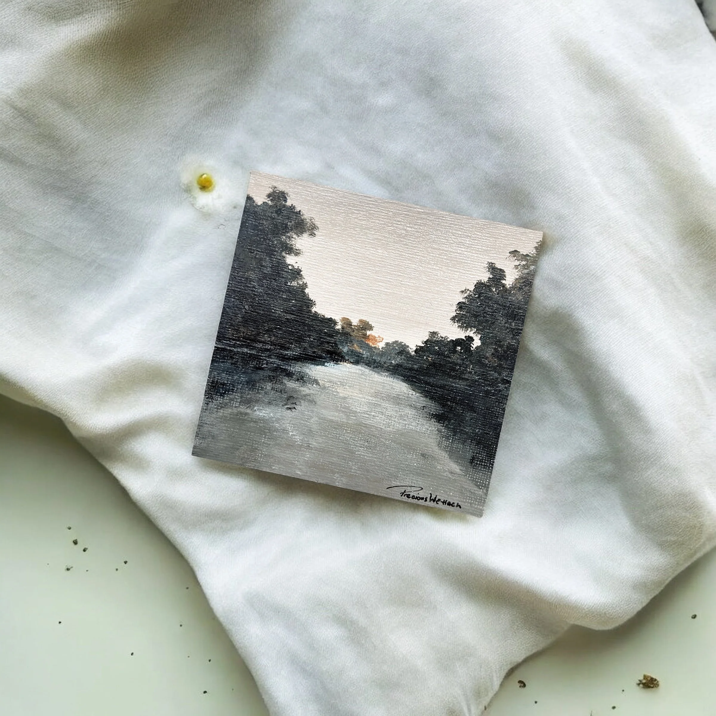 Small square landscape painting of a river surrounded by trees, signed by the artist, resting on white fabric with a small yellow and white flower nearby.