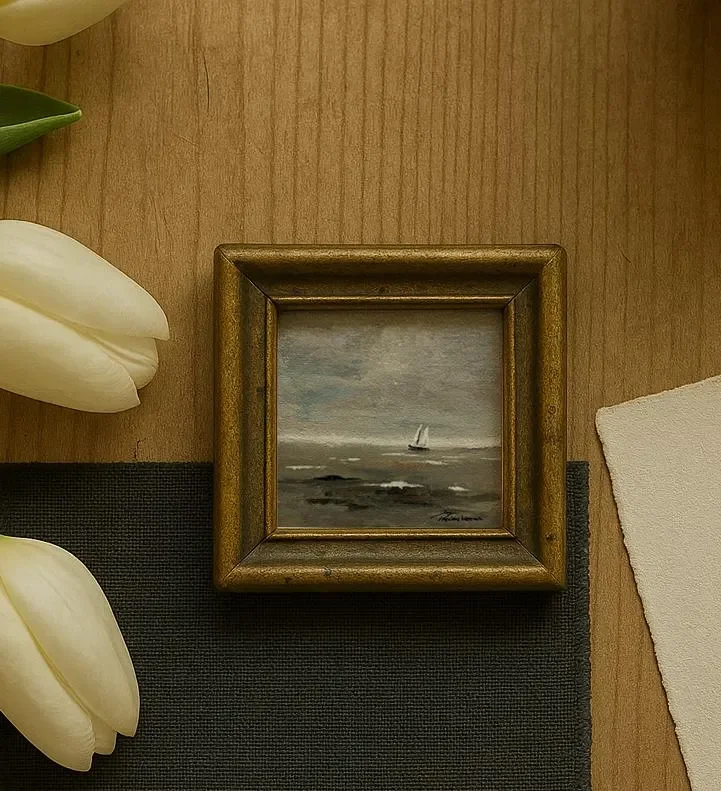 A small framed painting of a sailing boat on the ocean, placed on a wooden surface next to white tulips and a piece of off-white paper.