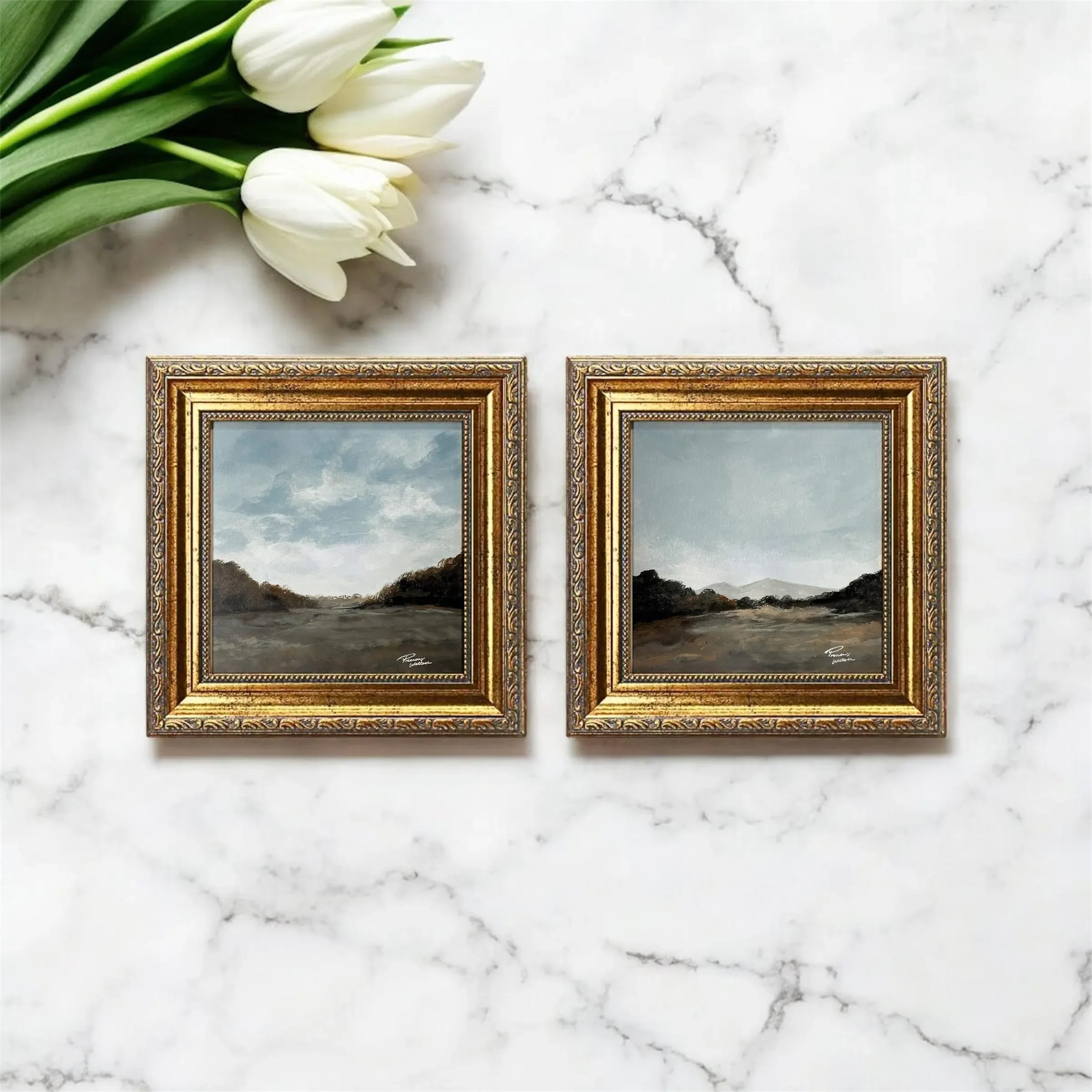 Two small landscape paintings in ornate golden frames placed on a white marble surface with a bouquet of white tulips nearby.