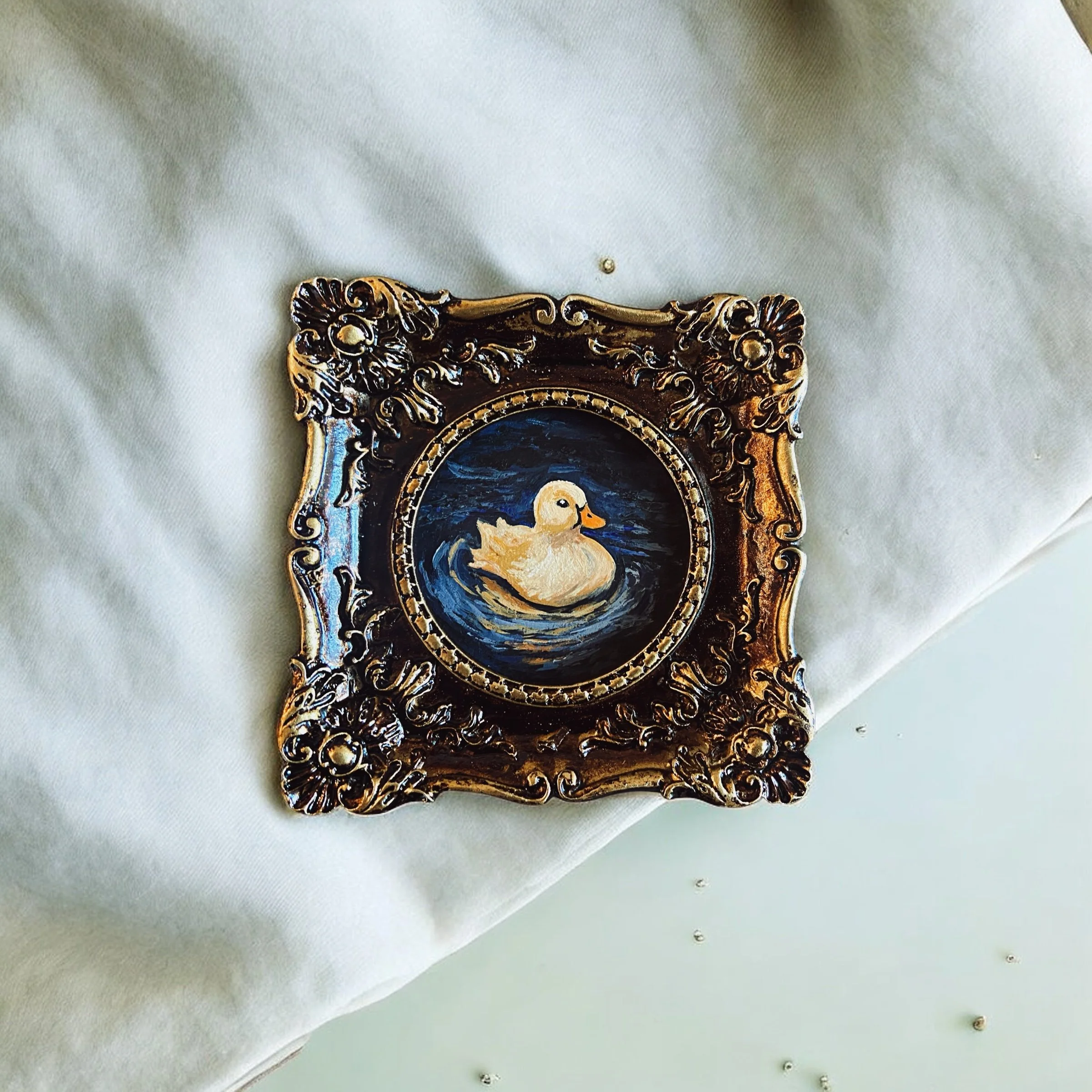 Decorative picture frame with an ornate gold design, containing a painting of a white duck swimming in blue water.