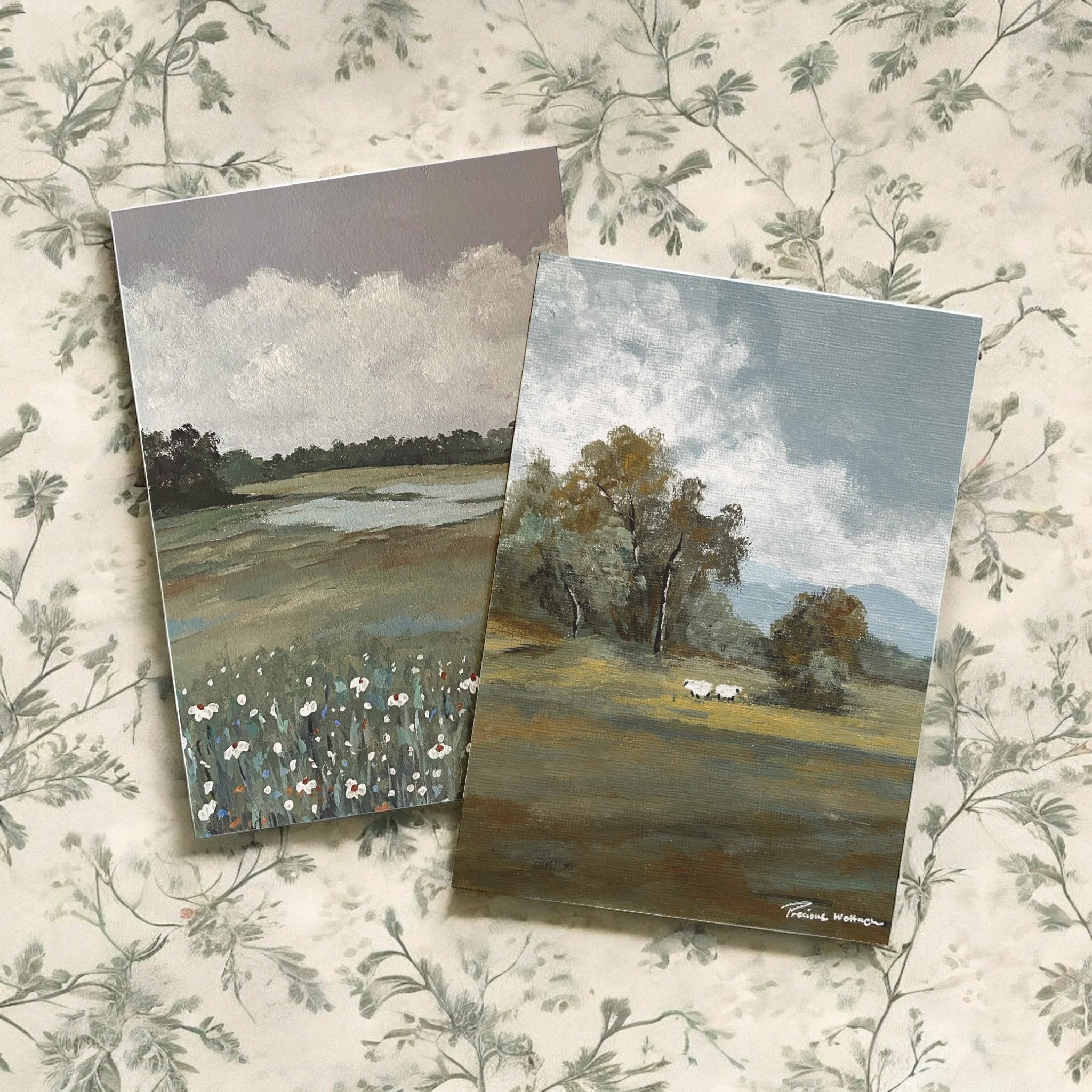 Two landscape paintings on paper featuring countryside scenes with trees, fields, clouds, and mountains, placed on a floral-patterned surface.