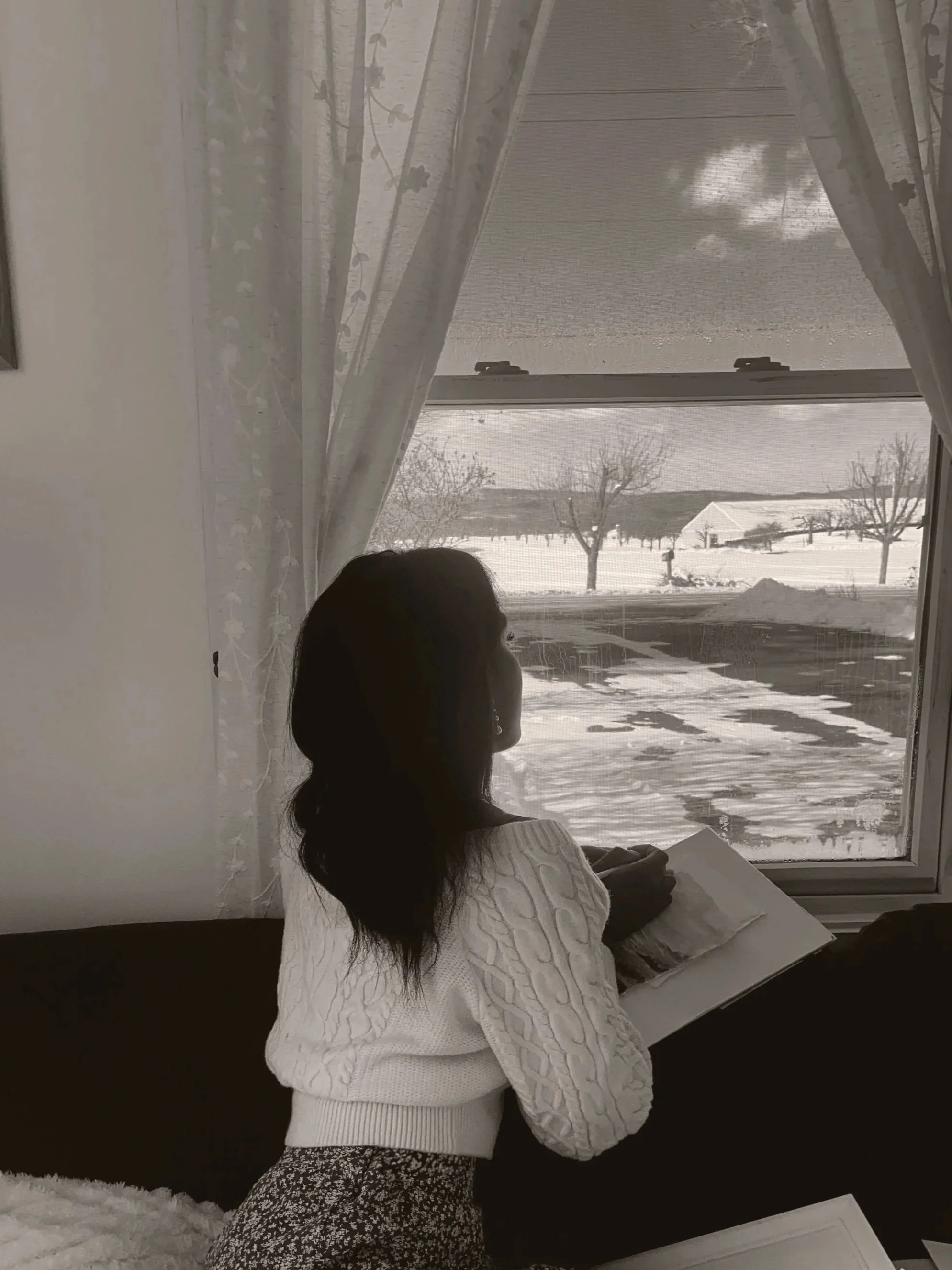 A woman with dark hair, wearing a white knit sweater and patterned pants, sits on a couch by a window, looking outside at snowy winter landscape with trees and houses.