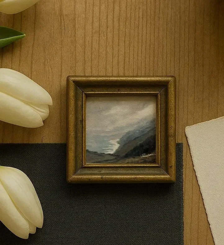 Small painting of a landscape with mountains, cloudy sky, and dark tones, framed in wood, resting on a wooden surface next to white tulips and a white piece of cloth.