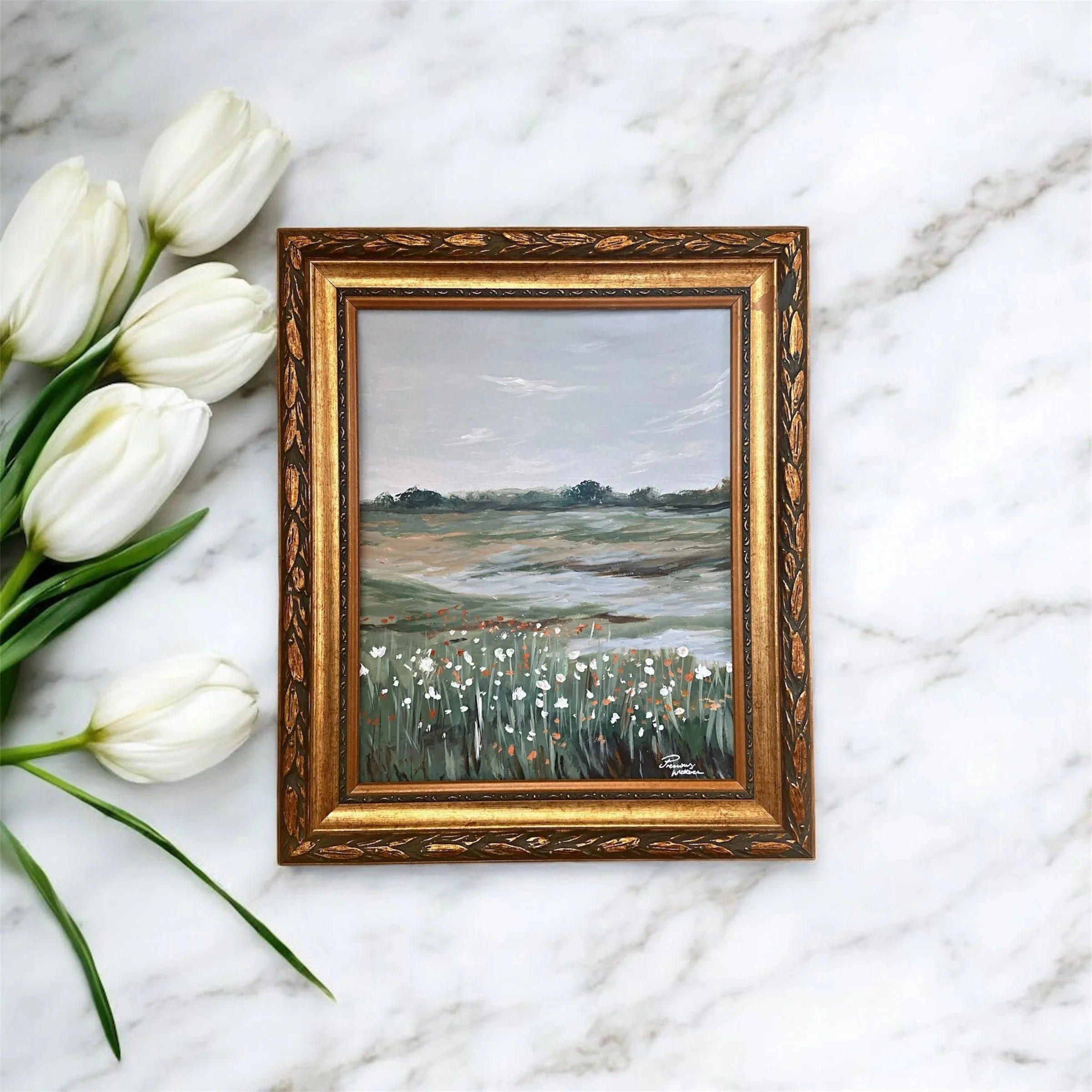A landscape painting of a cloudy sky, fields, and wildflowers in a decorative wooden orante frame, placed on a marble surface with white tulips on the side.
