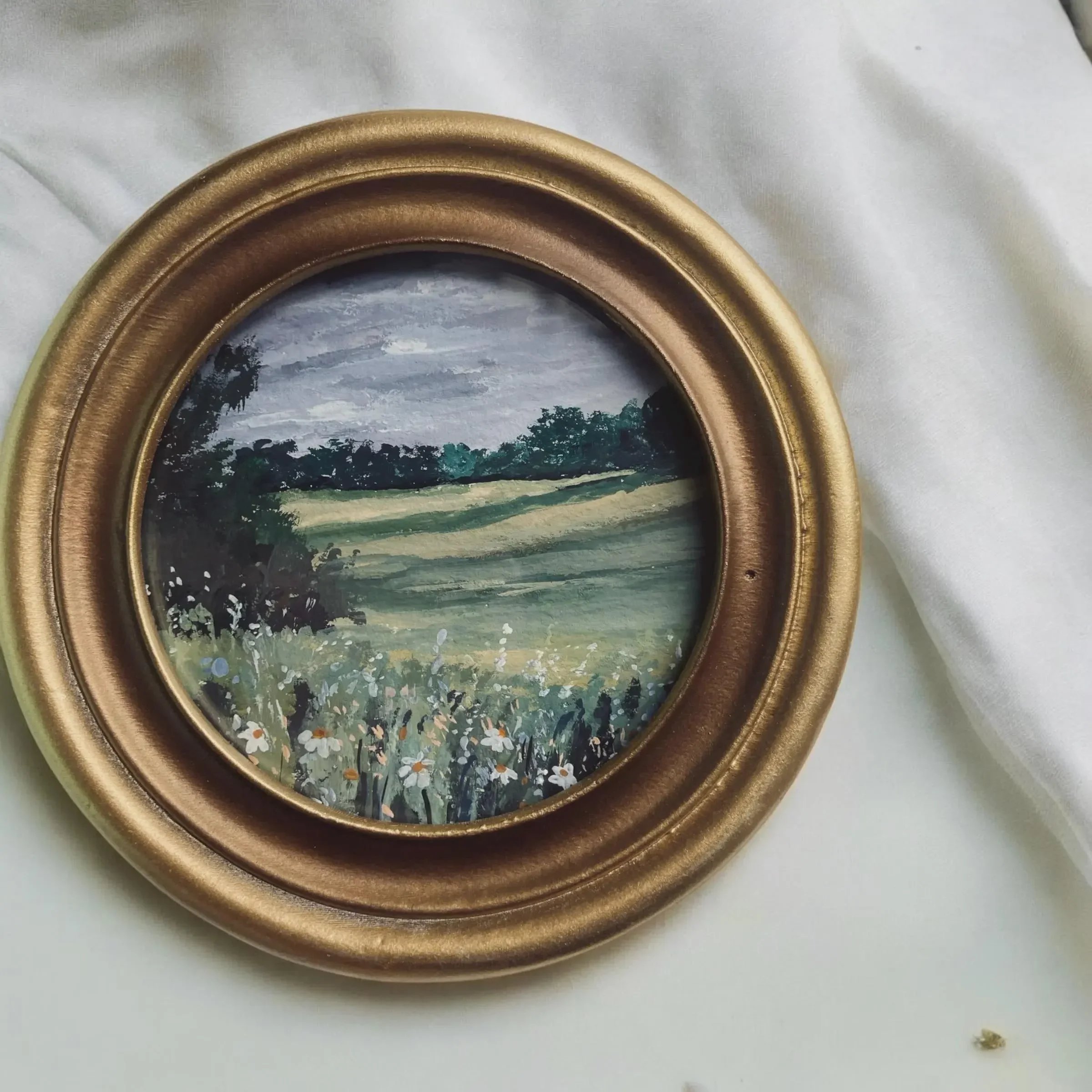 A small landscape painting of a field with flowers, trees, and cloudy sky, framed with a gold oval frame, placed on white fabric.