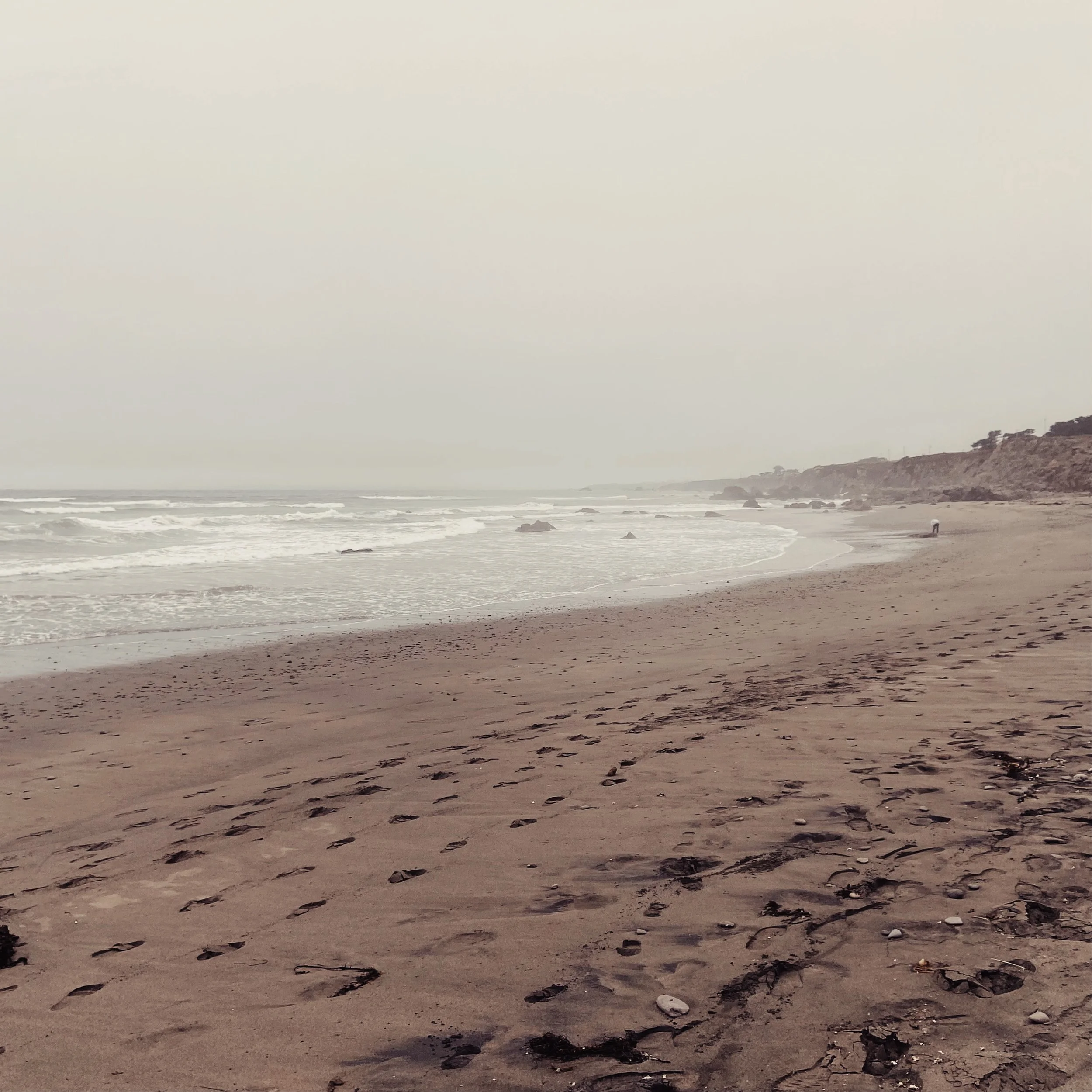 Overcast day at a sandy beach with footprints, waves, rocks, and distant cliffs.