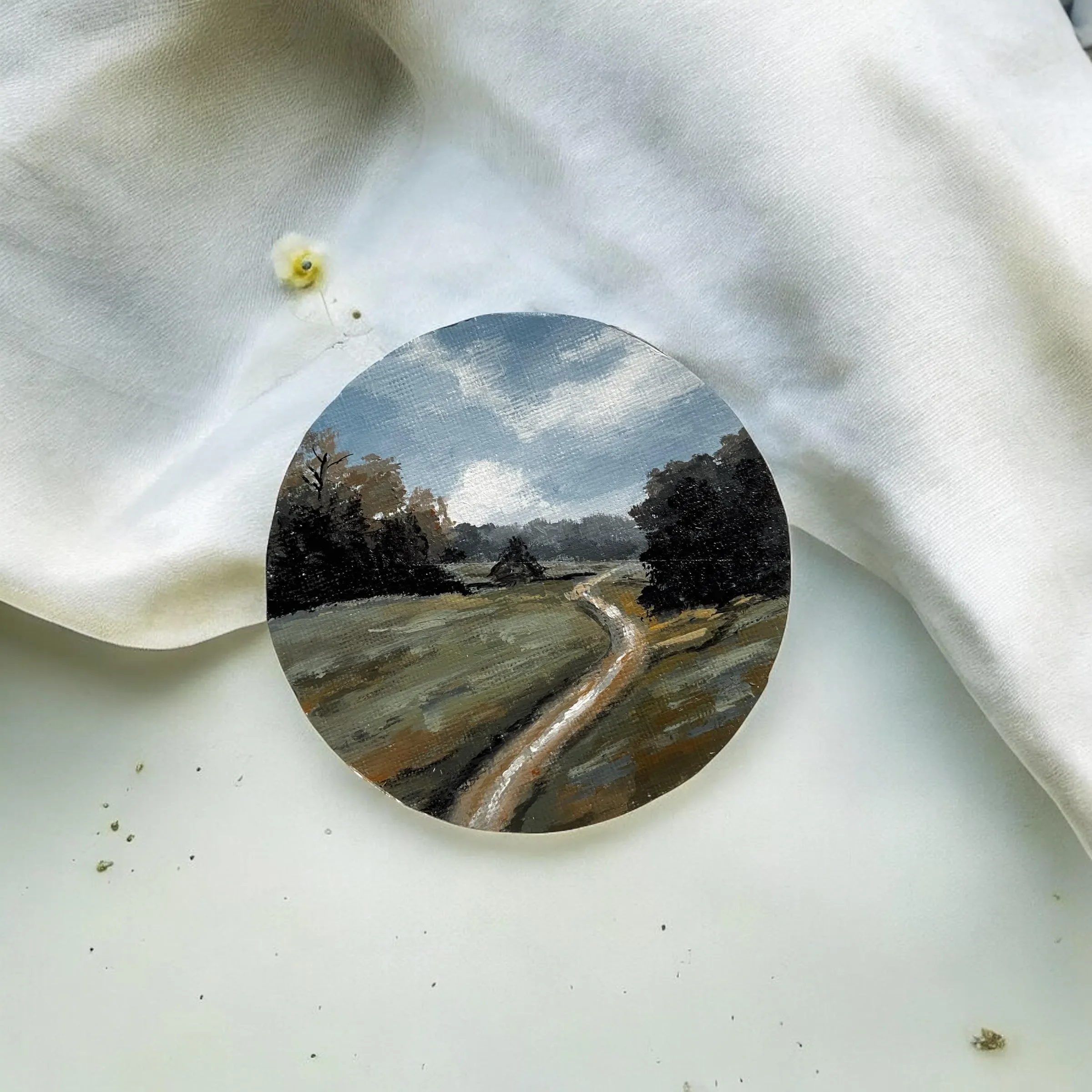 A circular painting of a rural landscape with a dirt path, trees, and a partly cloudy sky, placed on a white surface with a white fabric partially covering it.