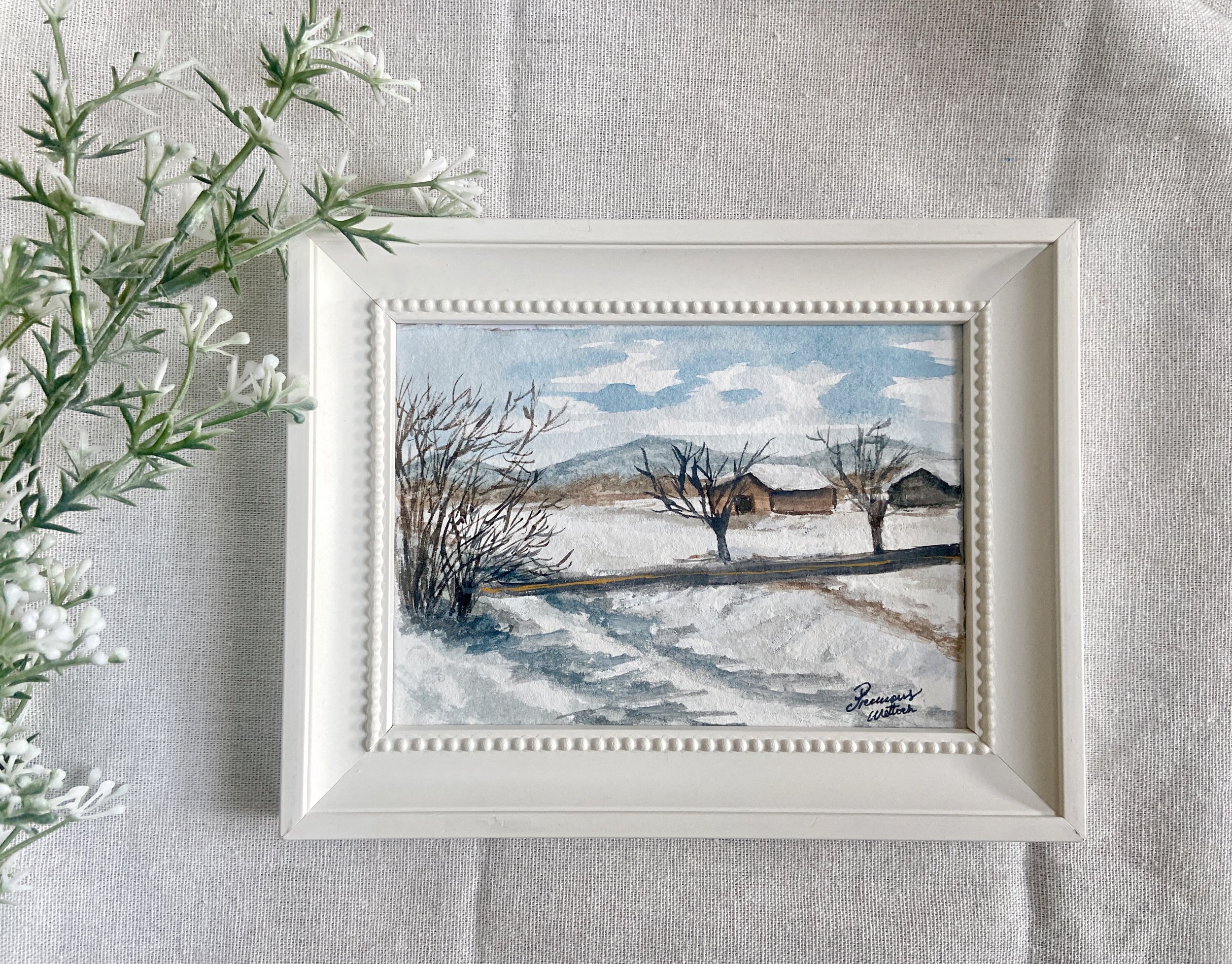 Watercolor painting of a winter landscape with snow-covered ground, leafless trees, and a few small houses, signed by the artist Precious Wettach.