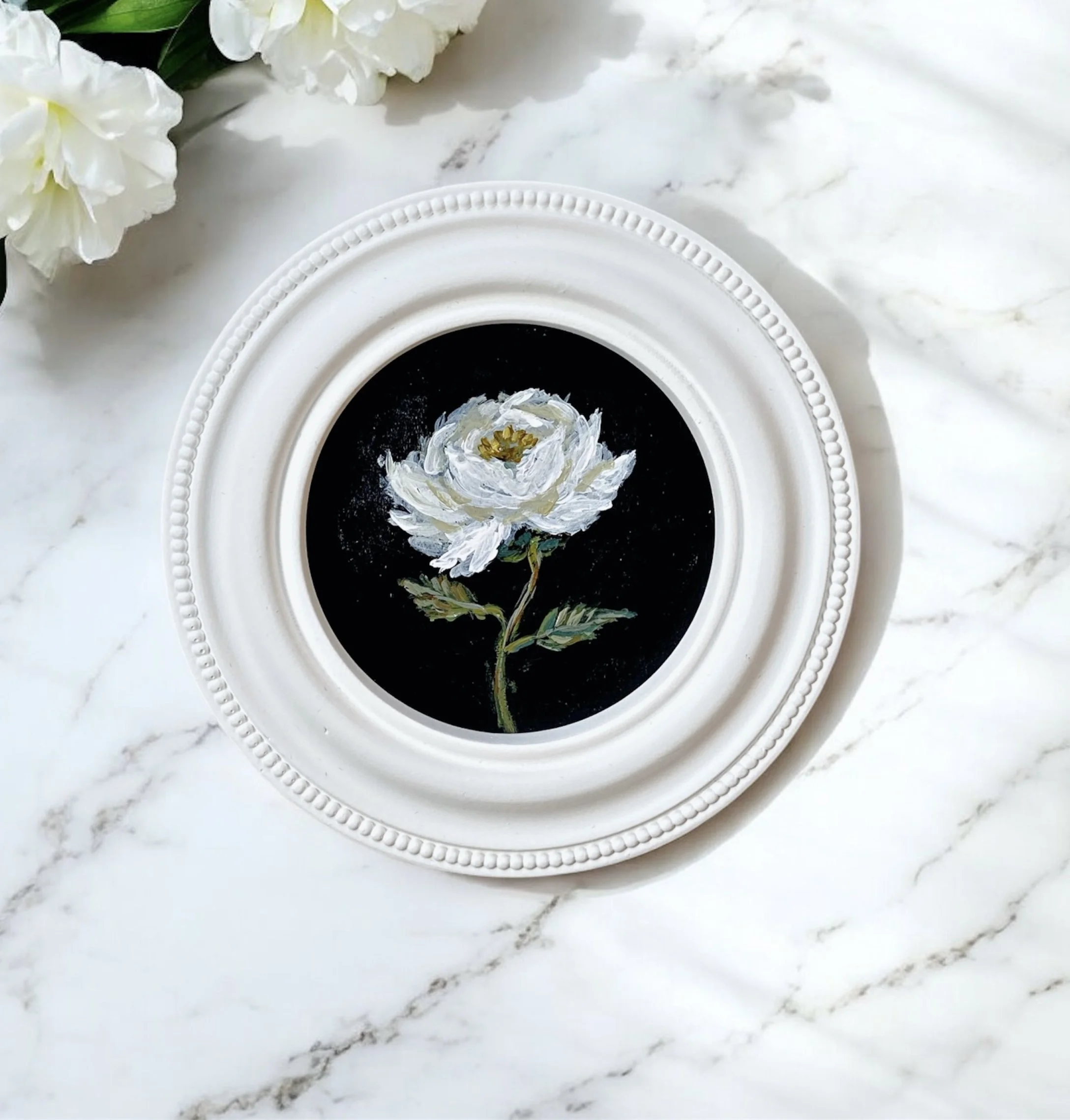 A framed painting of a white flower, placed on a white marble surface, with white flowers partially visible in the top left corner.