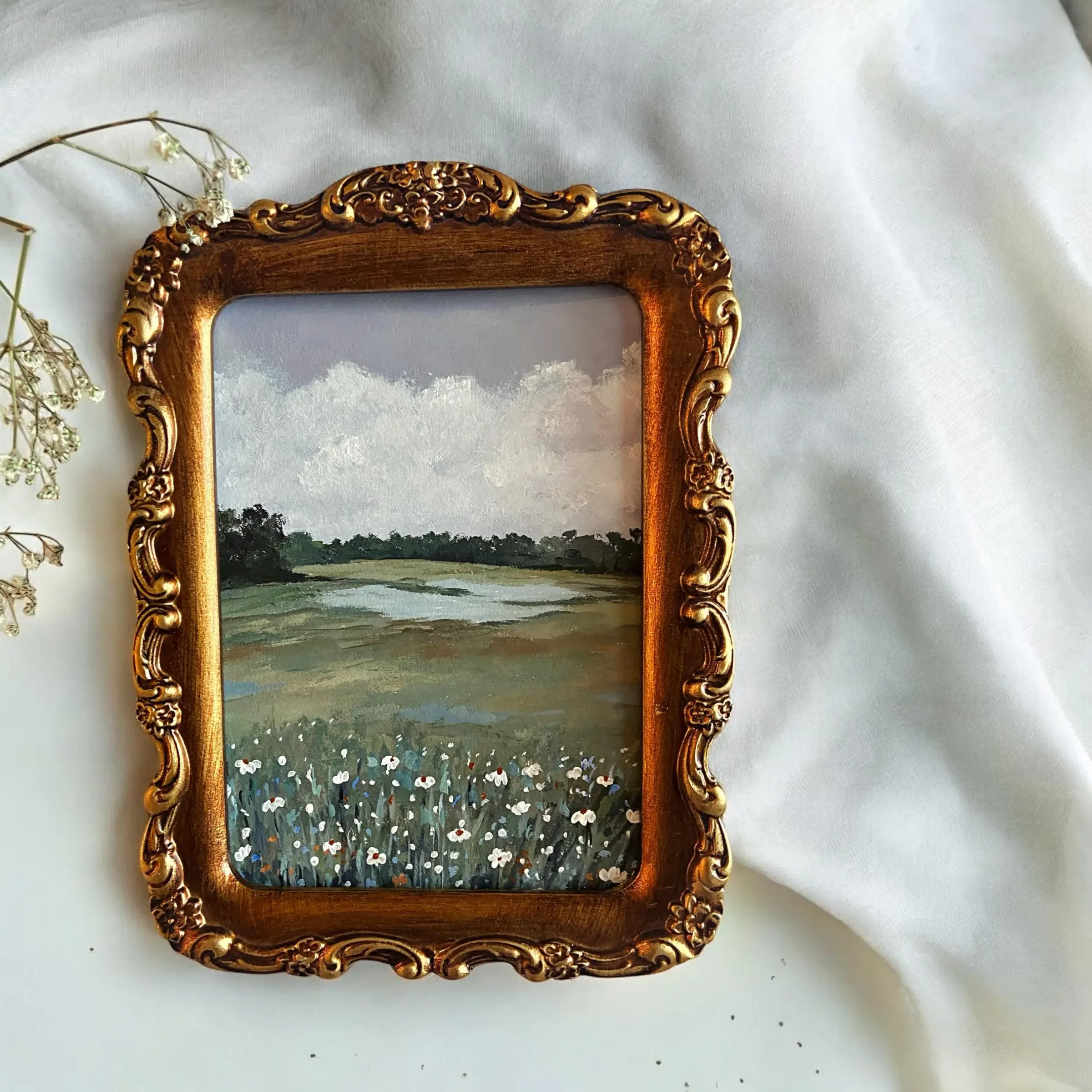 Small landscape painting of a field with flowers, trees, and cloudy sky, framed in an ornate gold picture frame, set on a white cloth with some dried flowers nearby.