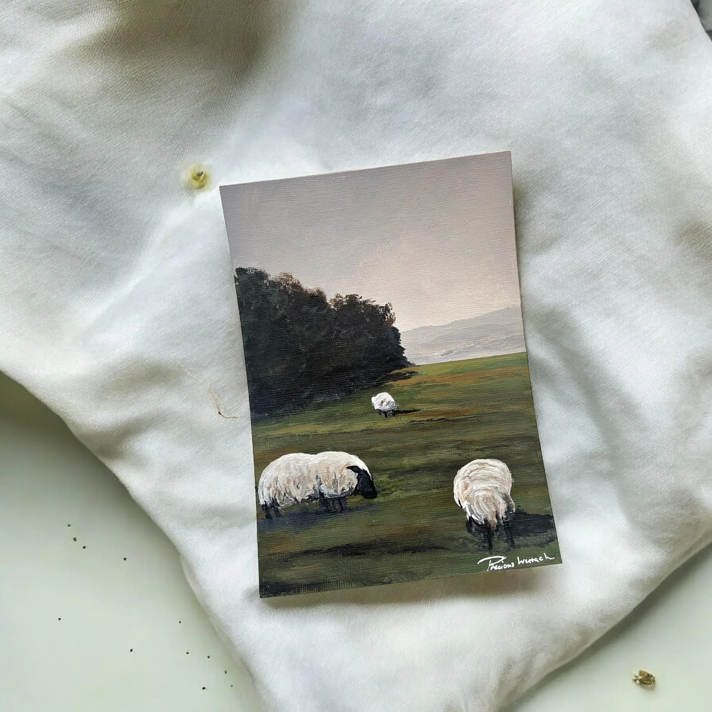 A small landscape painting depicting sheep grazing on green fields with trees and mountains in the background, signed by the artist in the bottom right corner.