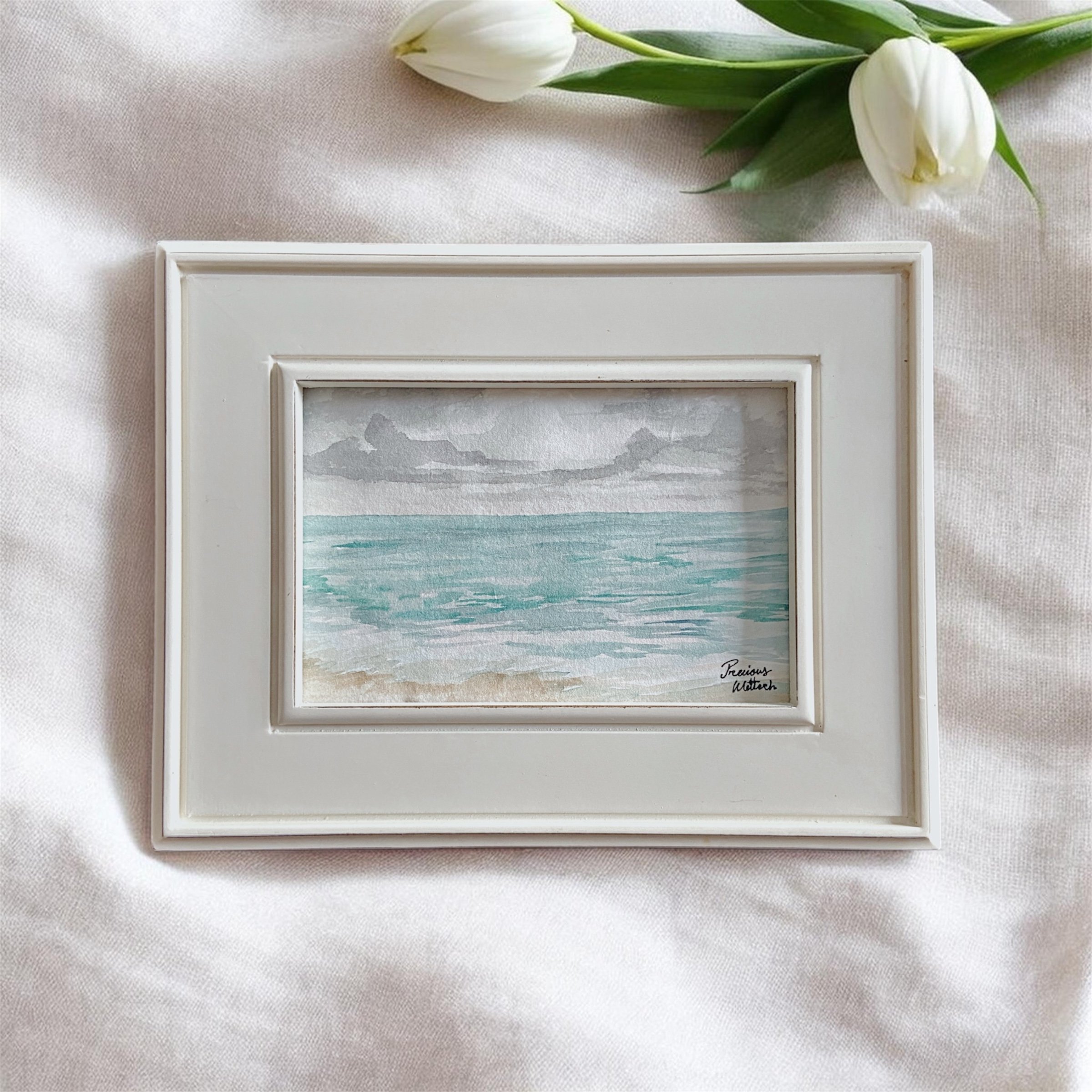 A framed watercolor painting of a seascape with grey clouds, turquoise water, and a sandy beach, placed on a white fabric surface with white tulips nearby.