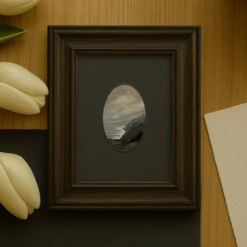 A framed painting of a cliffside seascape in a black oval mat, placed on a wooden surface next to cream-colored tulip-shaped objects and a white cloth.