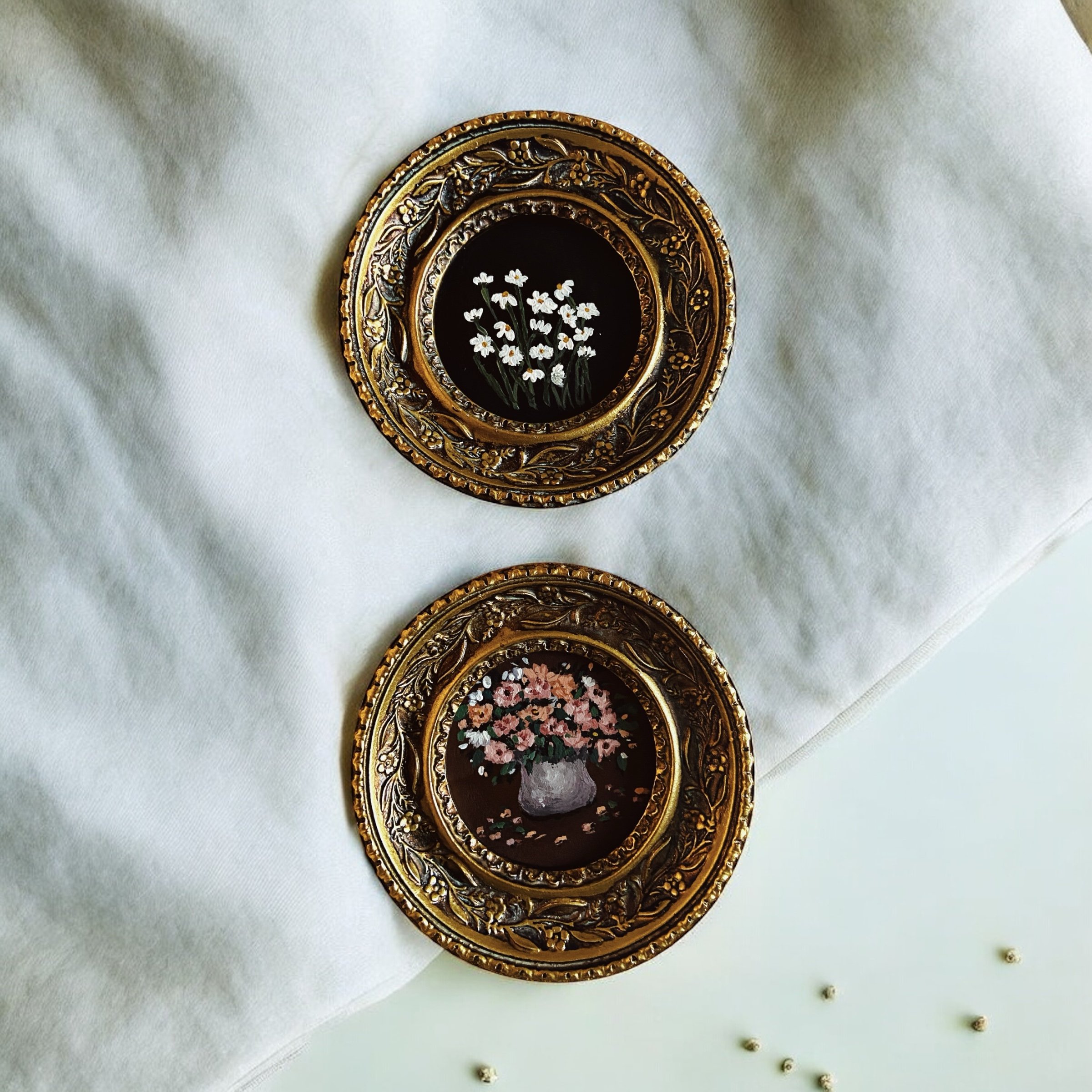 Two ornate gold picture frames with floral paintings placed on a white cloth surface.