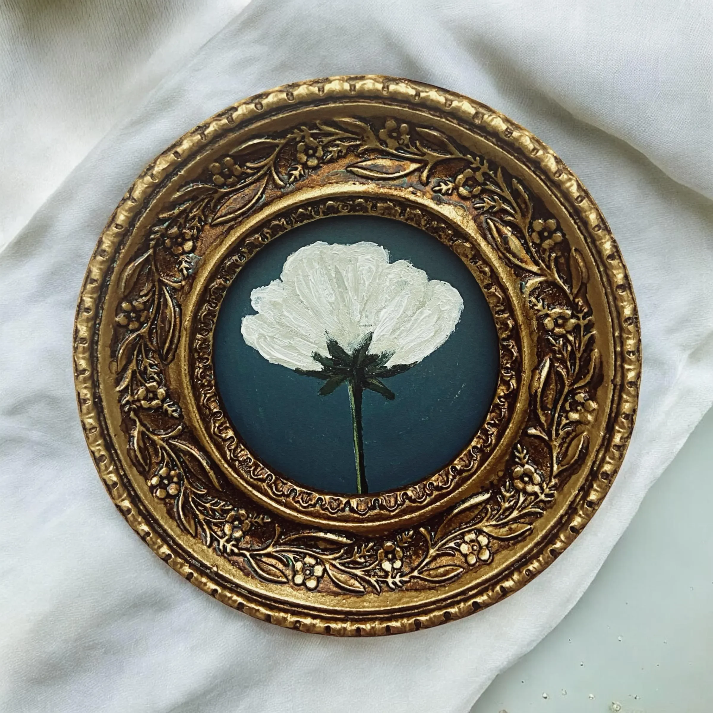 A gold ornate circular frame containing a painted white flower with a dark center and a green stem on a dark background.