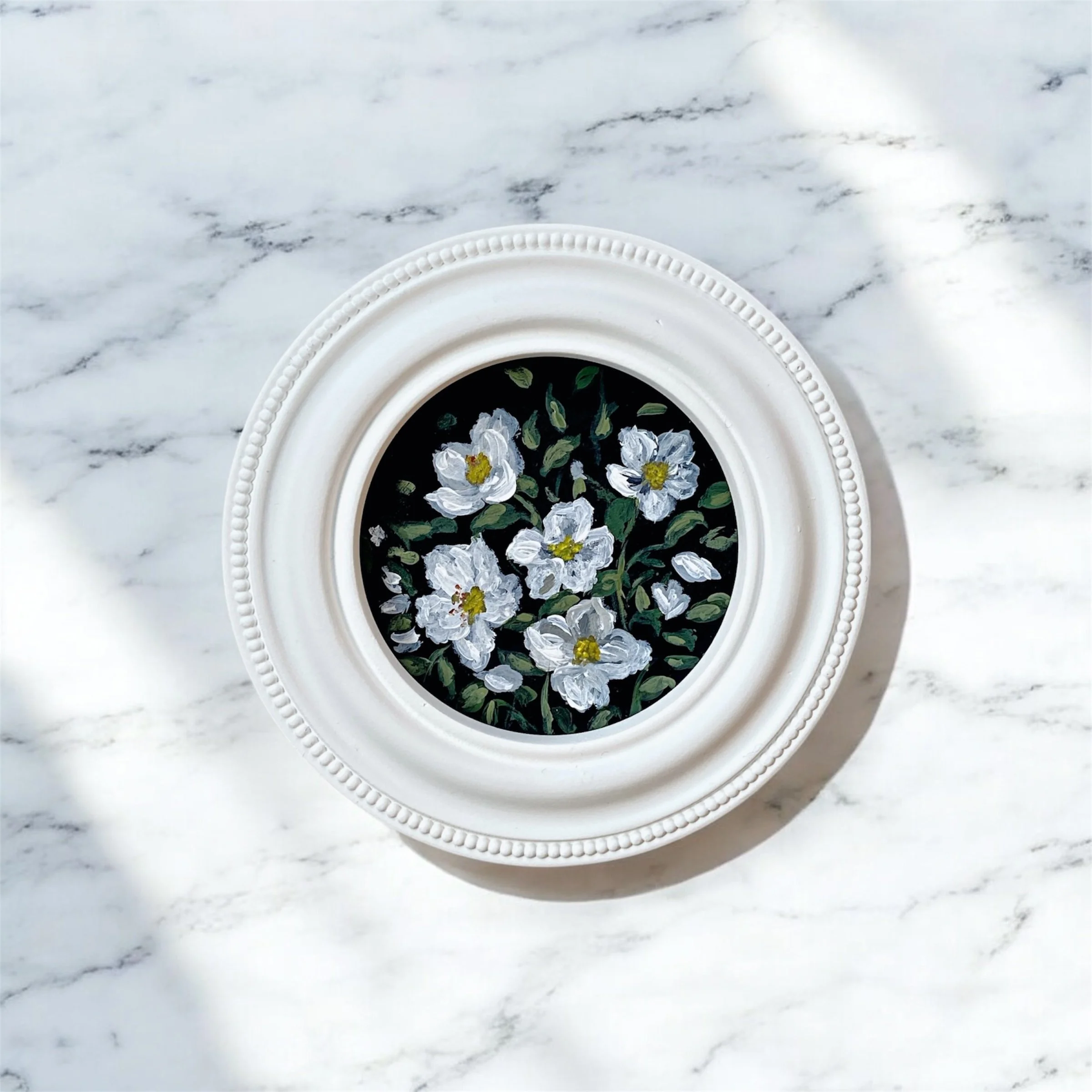 A round white picture frame with beaded detail, placed on a white marble surface, containing a painting of white flowers with green leaves on a black background.