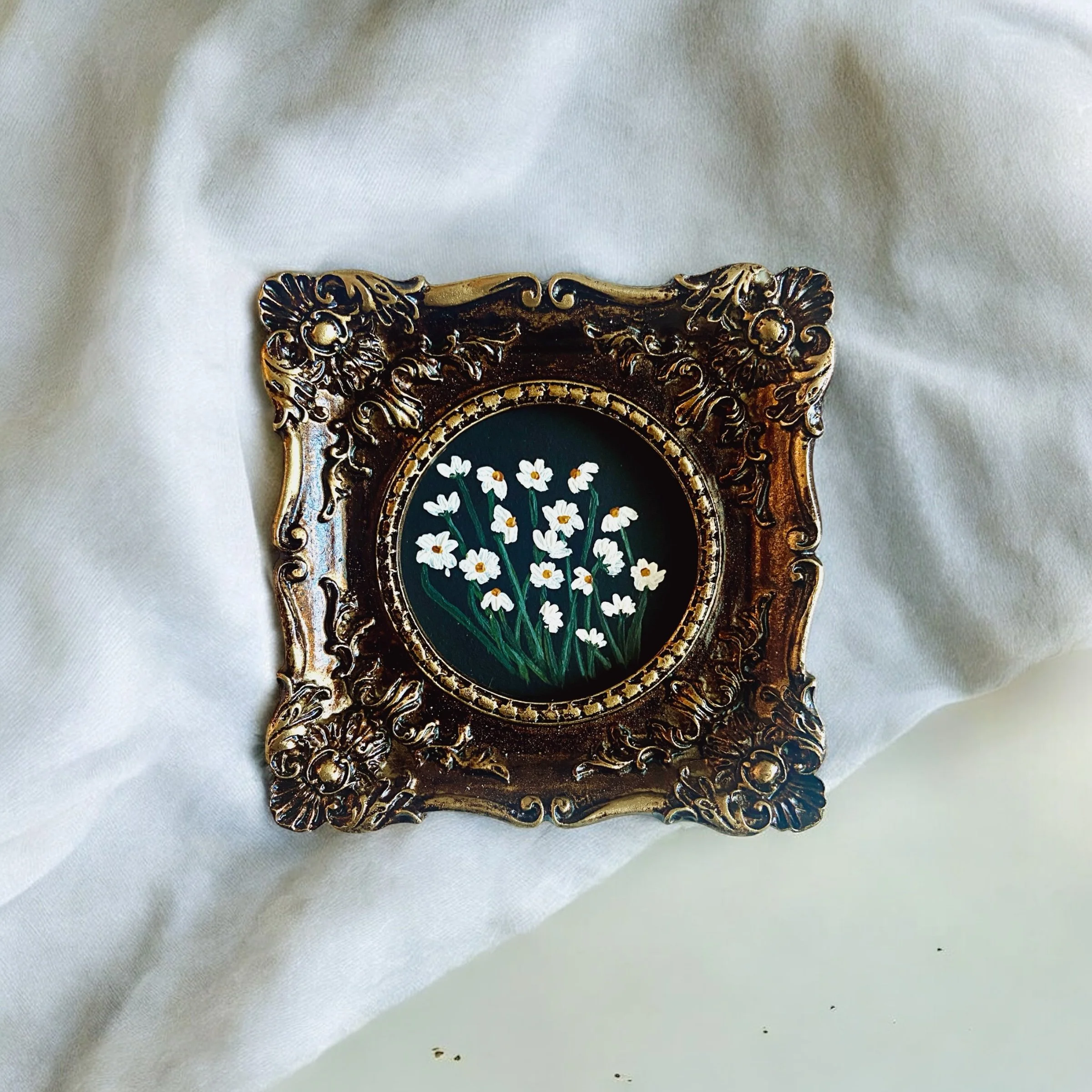 A decorative brass picture frame with ornate floral patterns, featuring a painting of white daisies on a dark background, placed on white fabric.