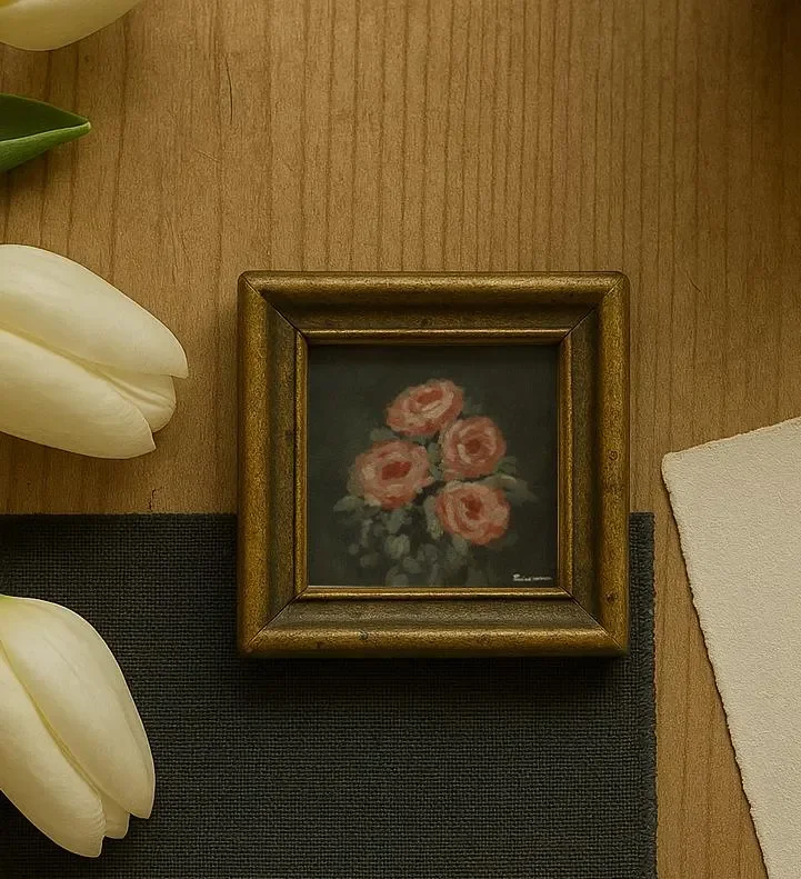 A small framed painting of pink roses on a dark background, placed on a wooden surface next to white tulips and a piece of torn white paper.