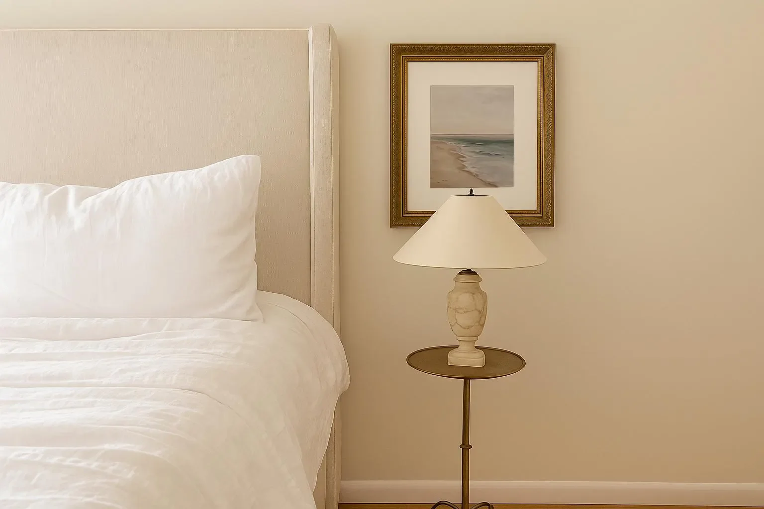 Close-up view of a neatly made bed with white pillows and bedding, next to a beige headboard, against a light-colored wall decorated with a framed seascape painting and a table lamp.