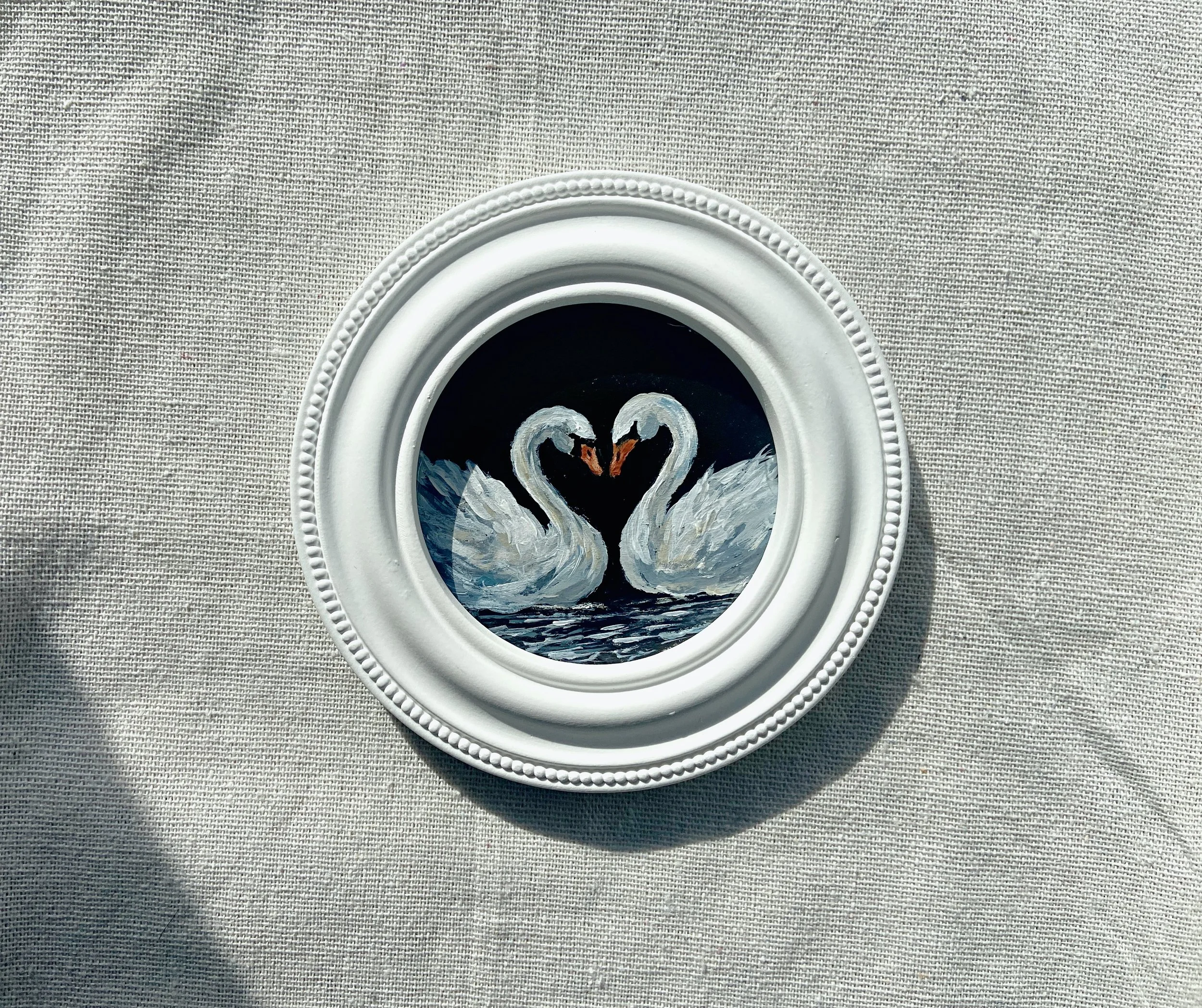 Painting of two swans touching beaks, framed in a white decorative frame, on a linen blanket.