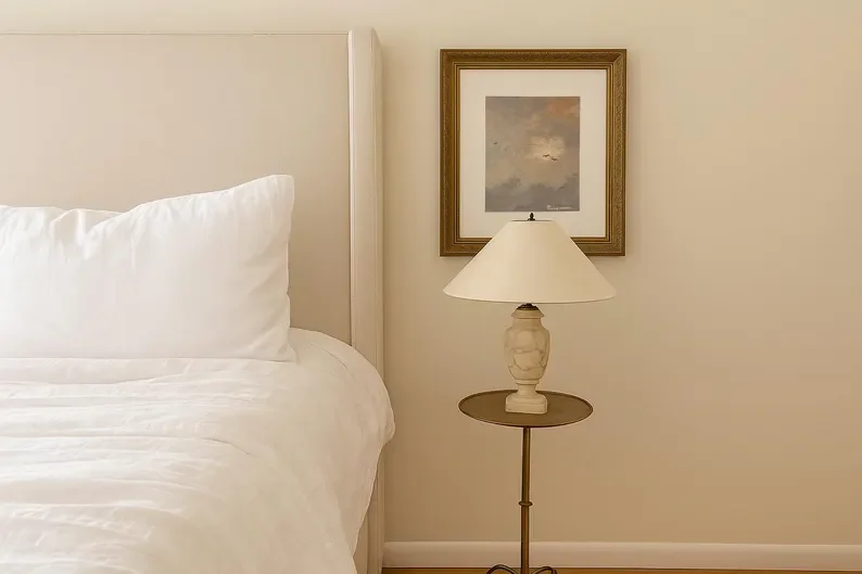 A bedroom with a white bed, a bedside table with a lamp, and framed soft painting of the sky on the wall.