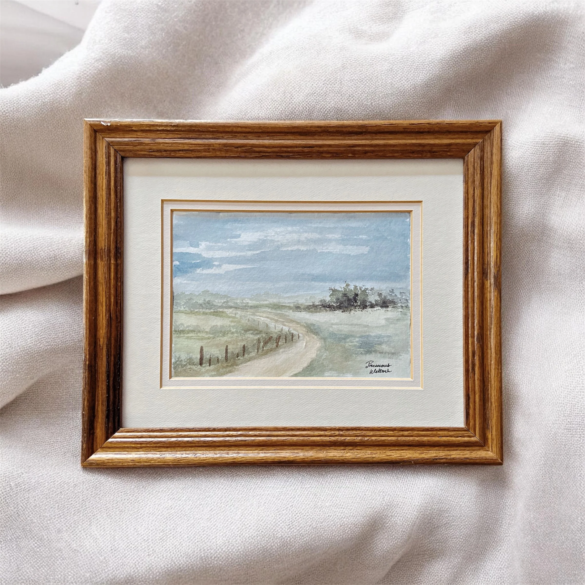 A framed watercolor painting of a rural landscape with a dirt road, grassy fields, trees, and a partly cloudy sky on white fabric.