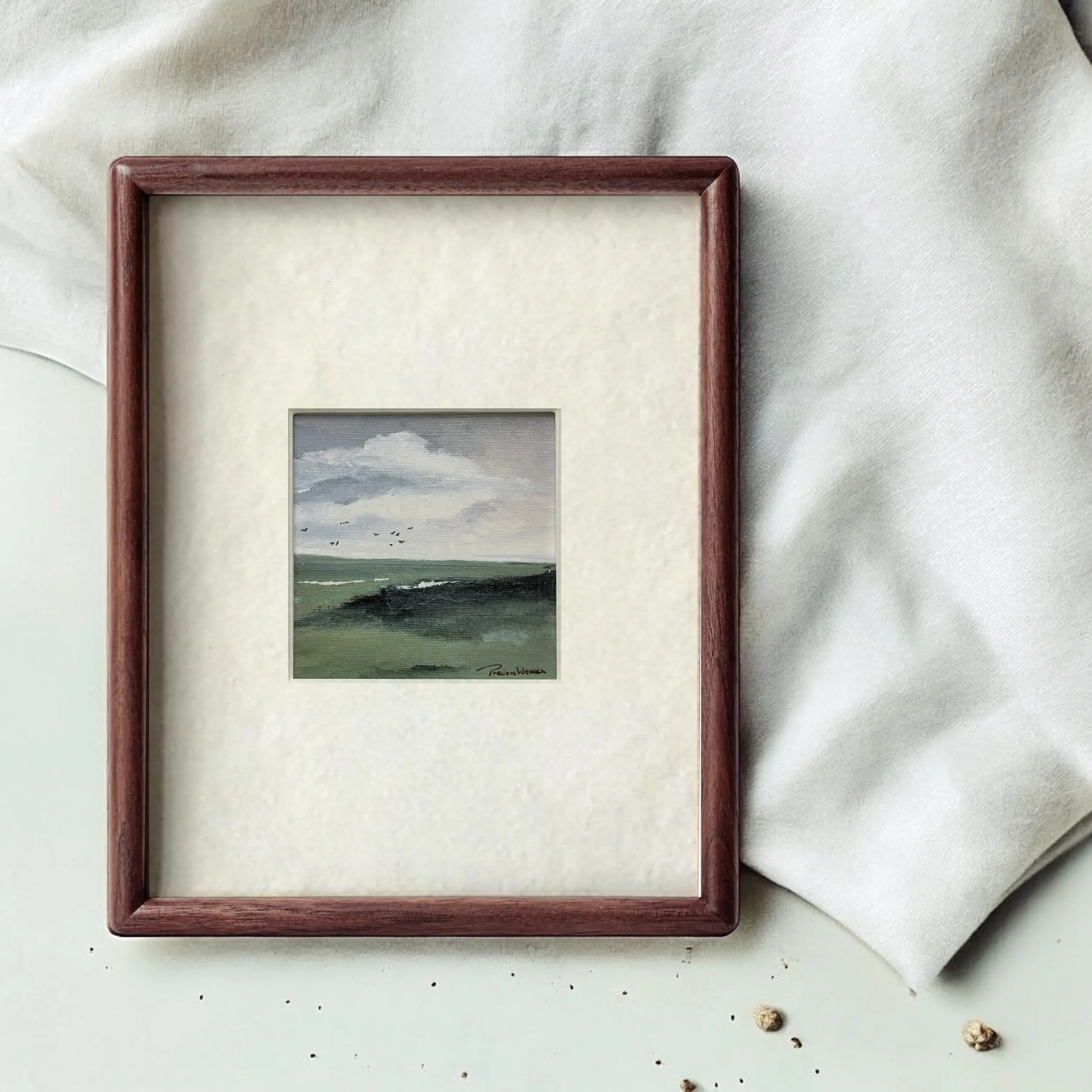 A framed miniature landscape painting of a cloudy sky, green fields, and distant birds on a table with white fabric and scattered small objects.