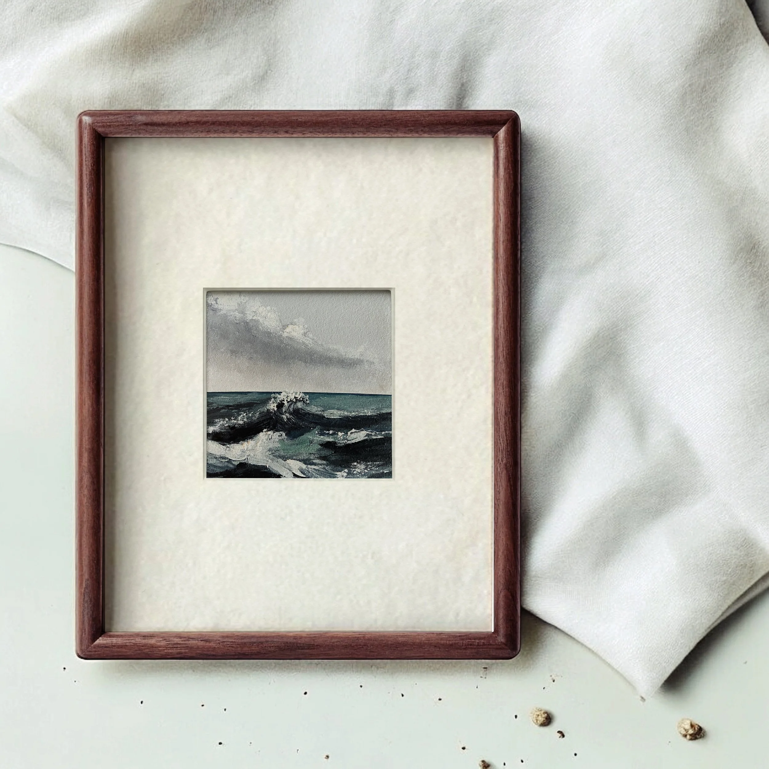 A framed miniature painting of a turbulent ocean with dark waves and a cloudy sky, placed on a soft white surface with some scattered small pebbles.