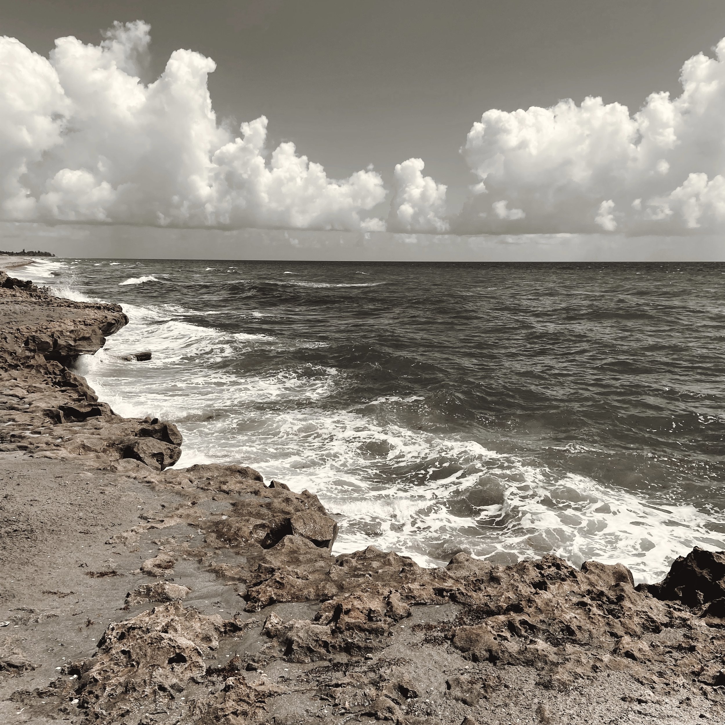 View of a rocky shoreline with waves crashing and an overcast sky with fluffy clouds above the ocean.