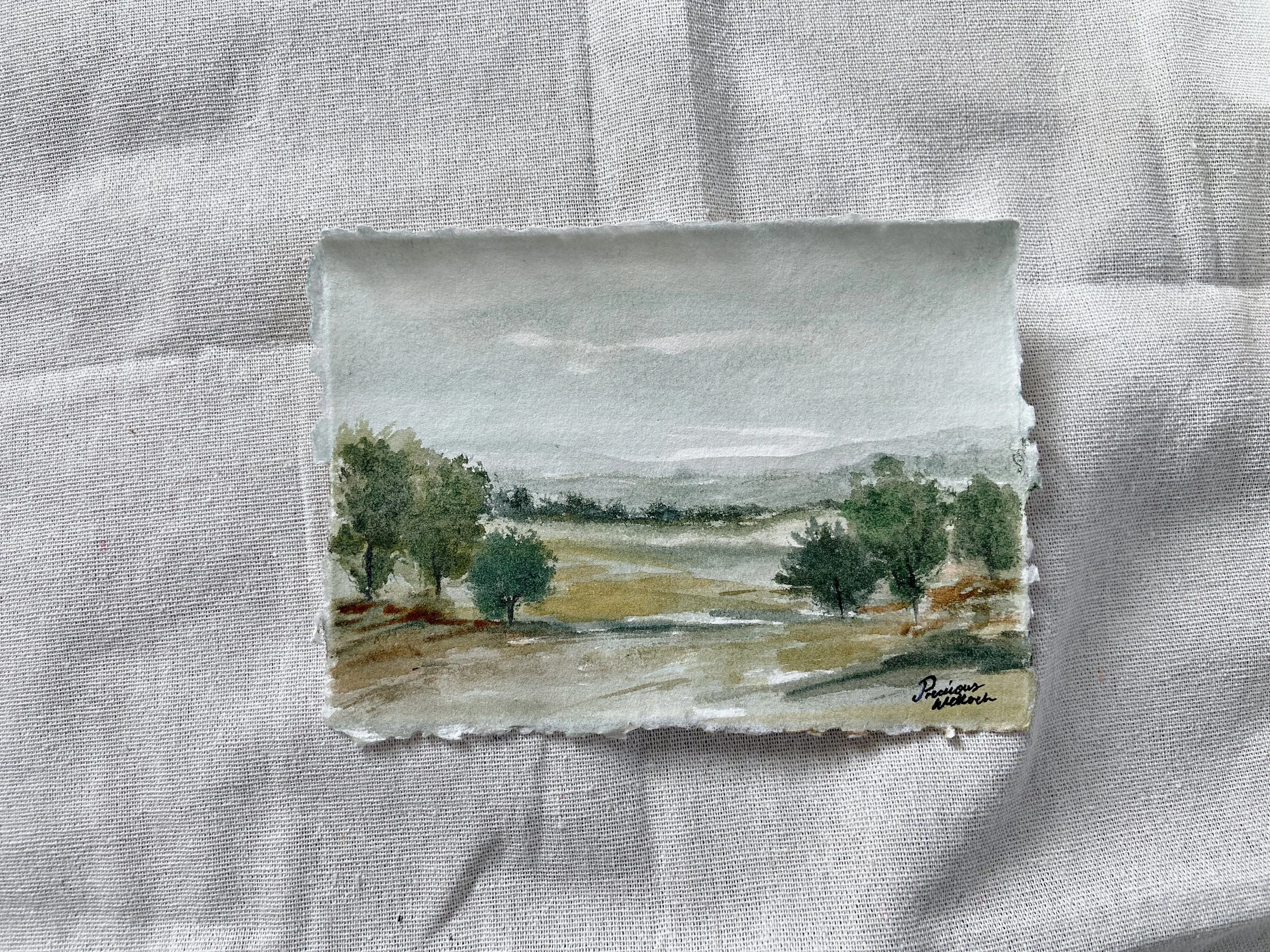 A small watercolor painting of a landscape with trees, a river, and rolling hills under a cloudy sky, signed in the lower right corner.