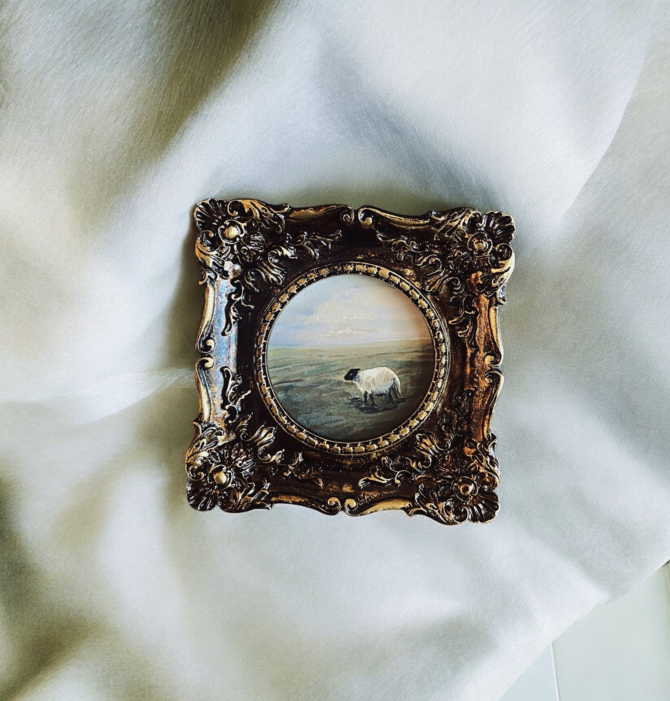 Decorative picture frame with a landscape painting of a sheep in a field, resting on white fabric.