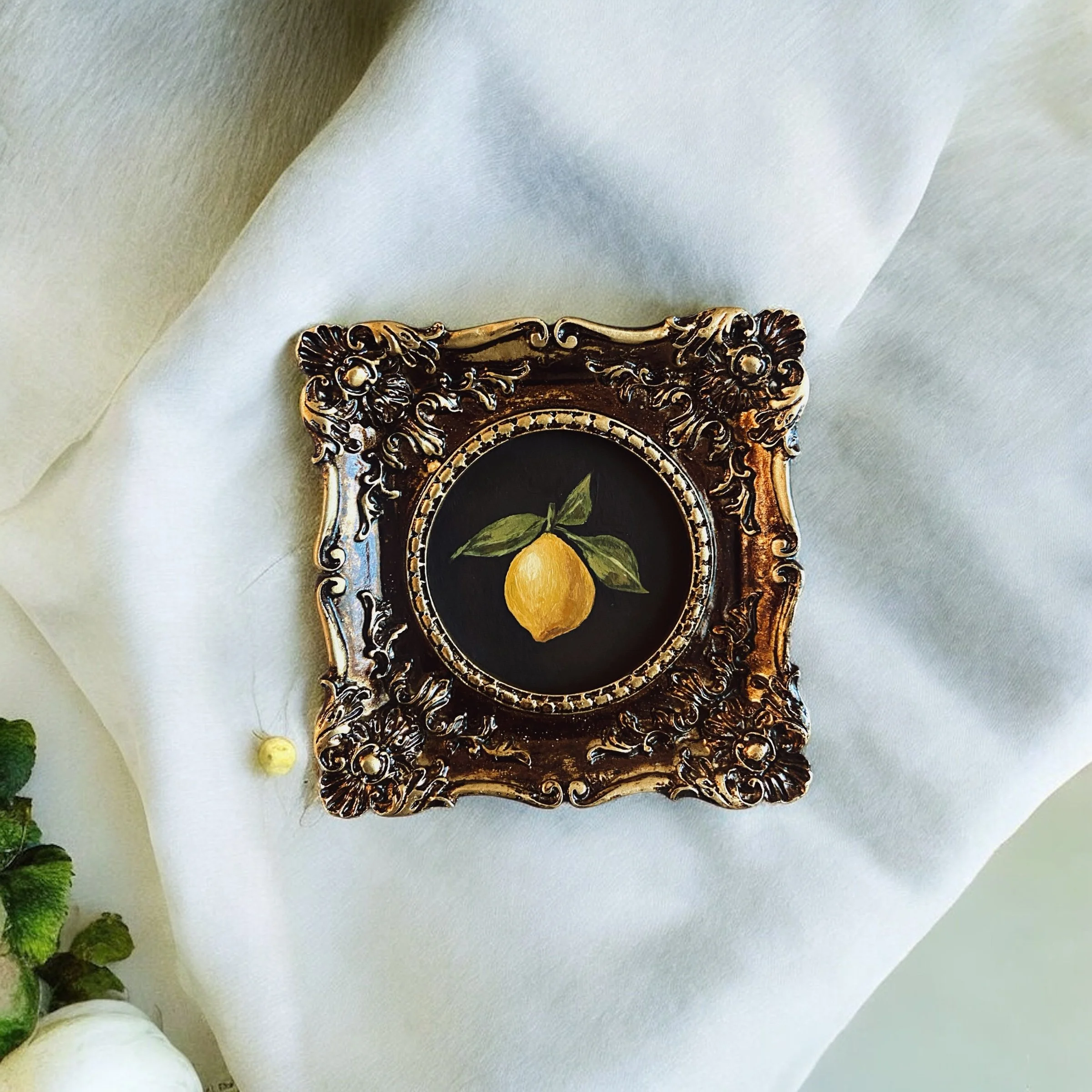 Ornate gold frame with a painted lemon and leaves, placed on white fabric.
