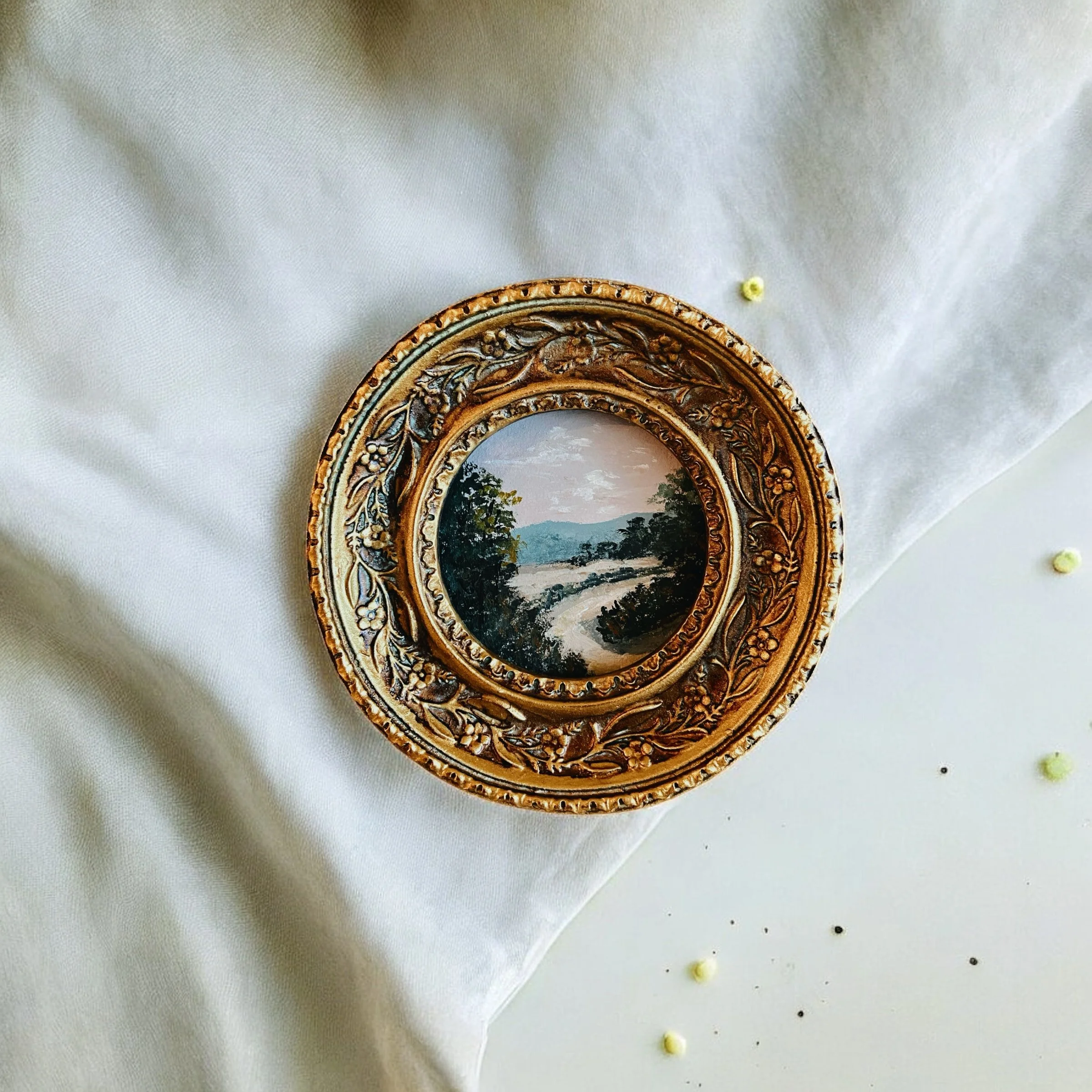 Decorative golden frame with a small oval landscape painting of a river, trees, and mountains, placed on a white fabric surface with scattered small yellow and black beads.