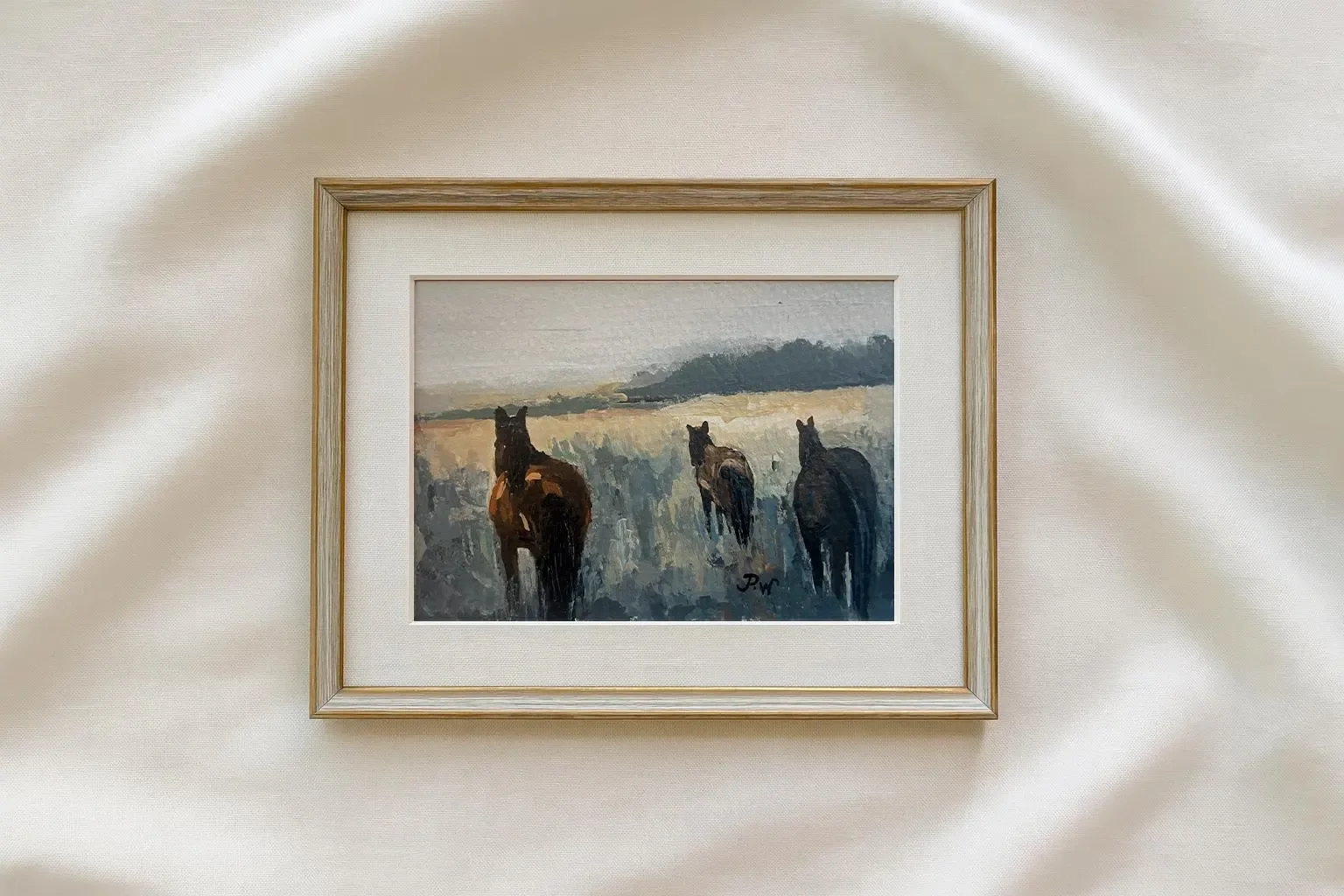 Framed watercolor painting of three horses running in an open field with distant trees and sky.