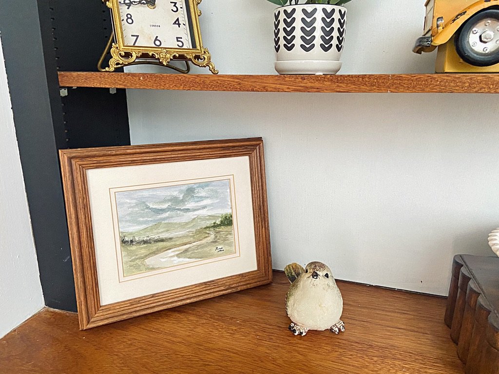 A framed landscape painting leaning against a wall, a small bird figurine on the wooden surface, and a decorative pot with a plant on the shelf above.